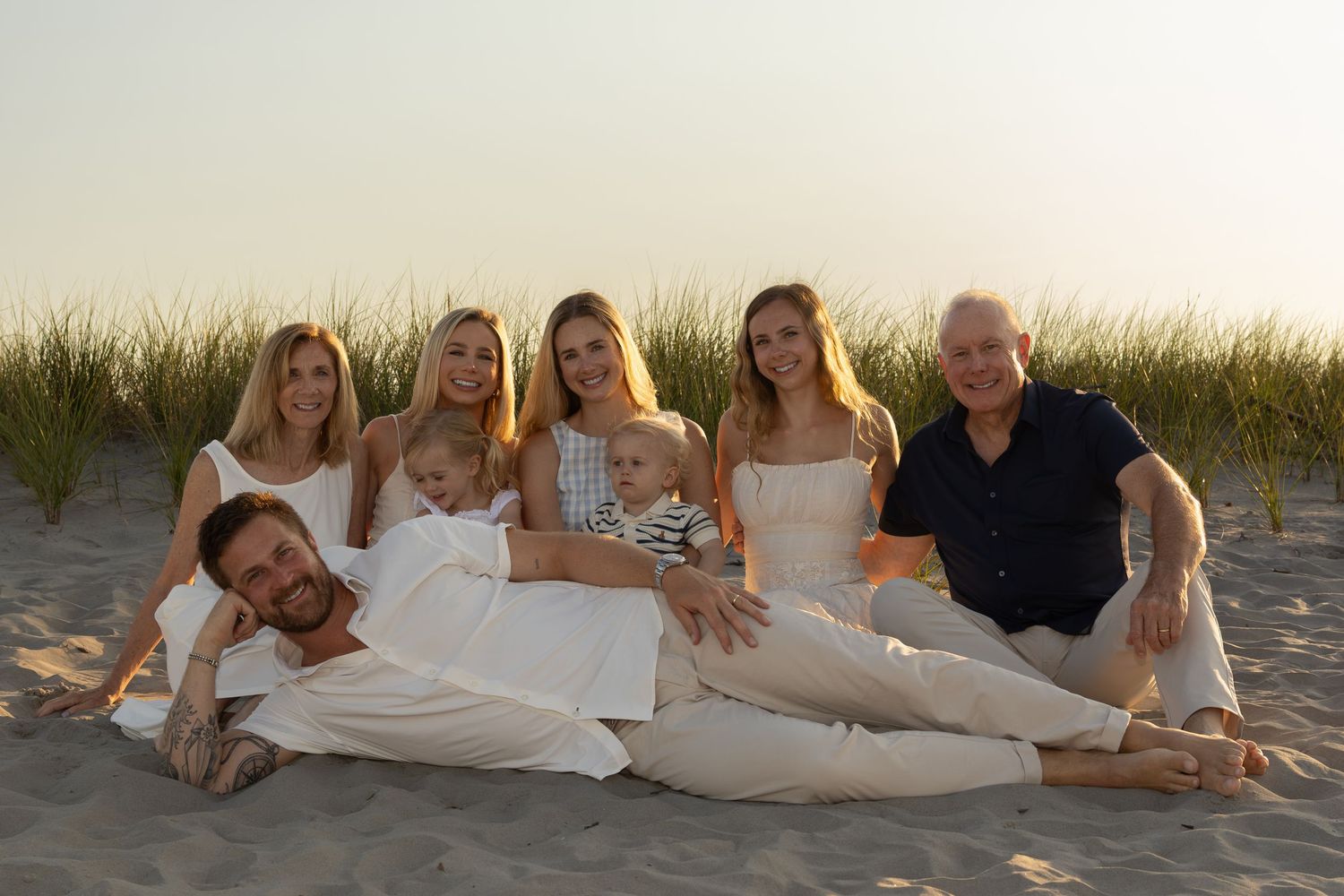 Large family group photo on sandy beach at sunset, adults and children in white outfits, smiling together.