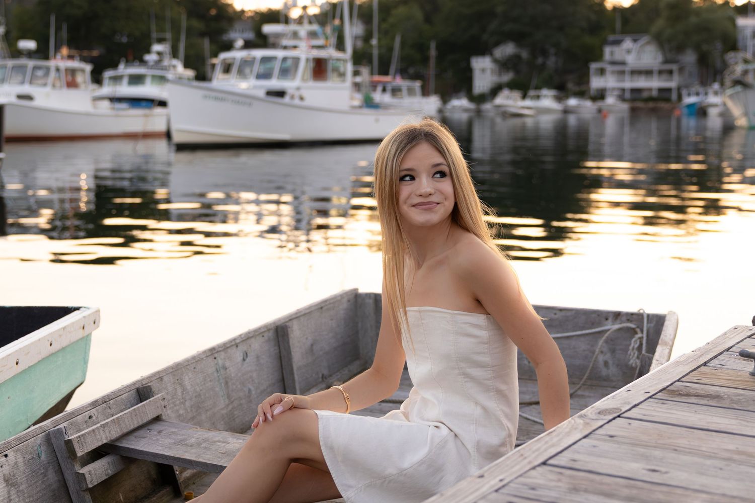 Young blonde woman in white dress sitting in a wooden boat at a marina during golden hour sunset.