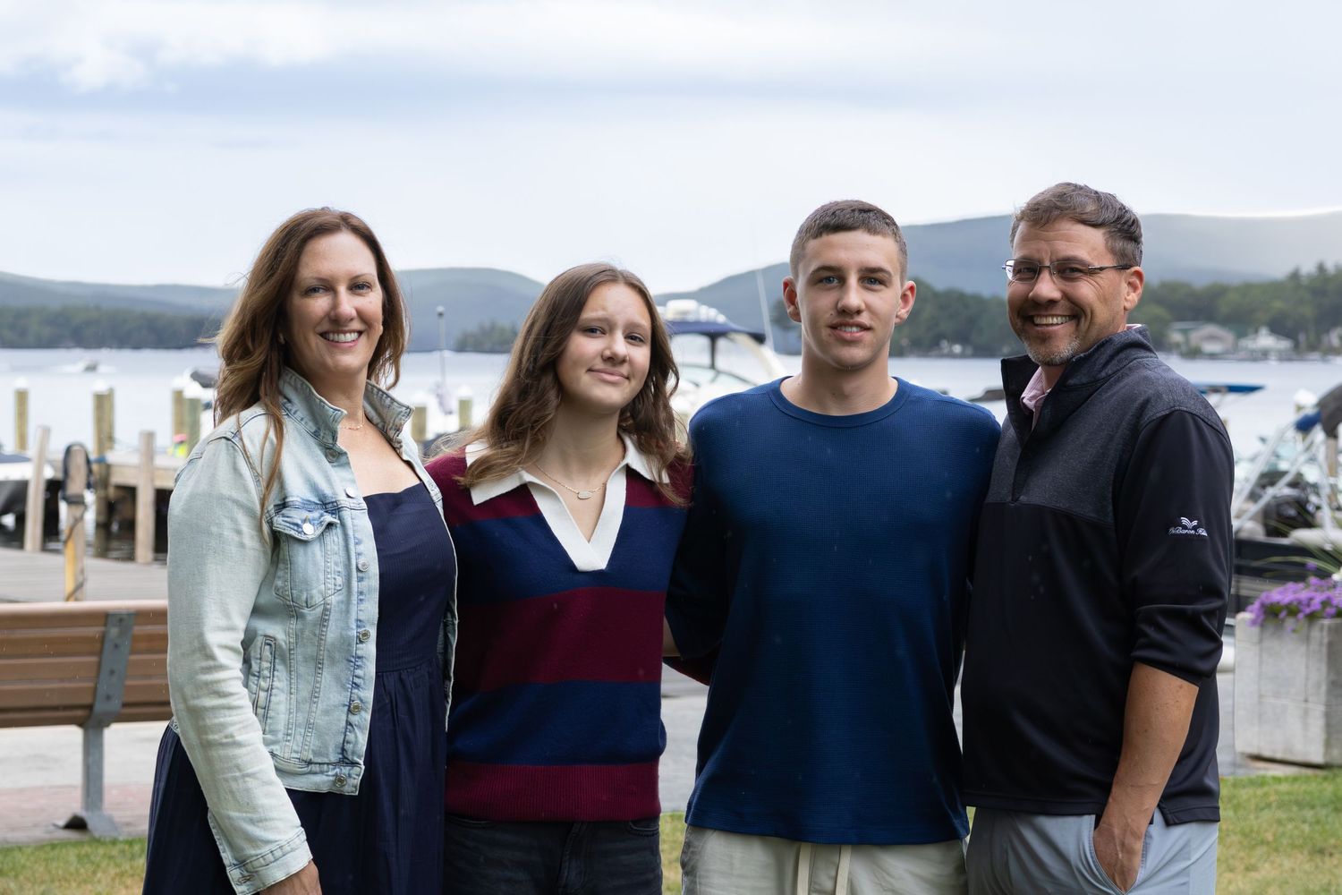 Family of four smiling outdoors near a lake with mountains in the background on a cloudy day.