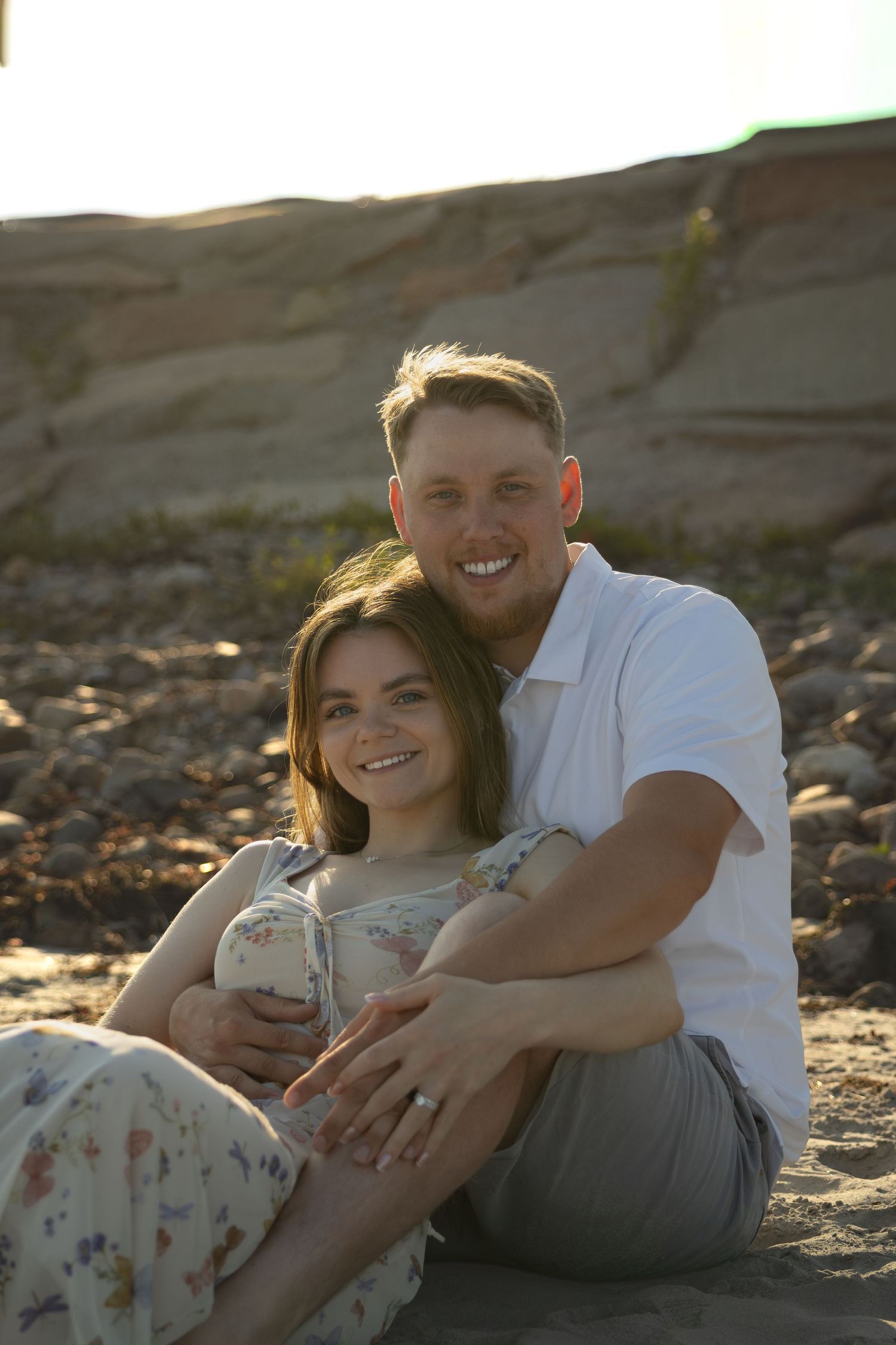 A smiling couple sits together on a sandy beach, the man in white embracing the woman in a floral dress at golden hour.