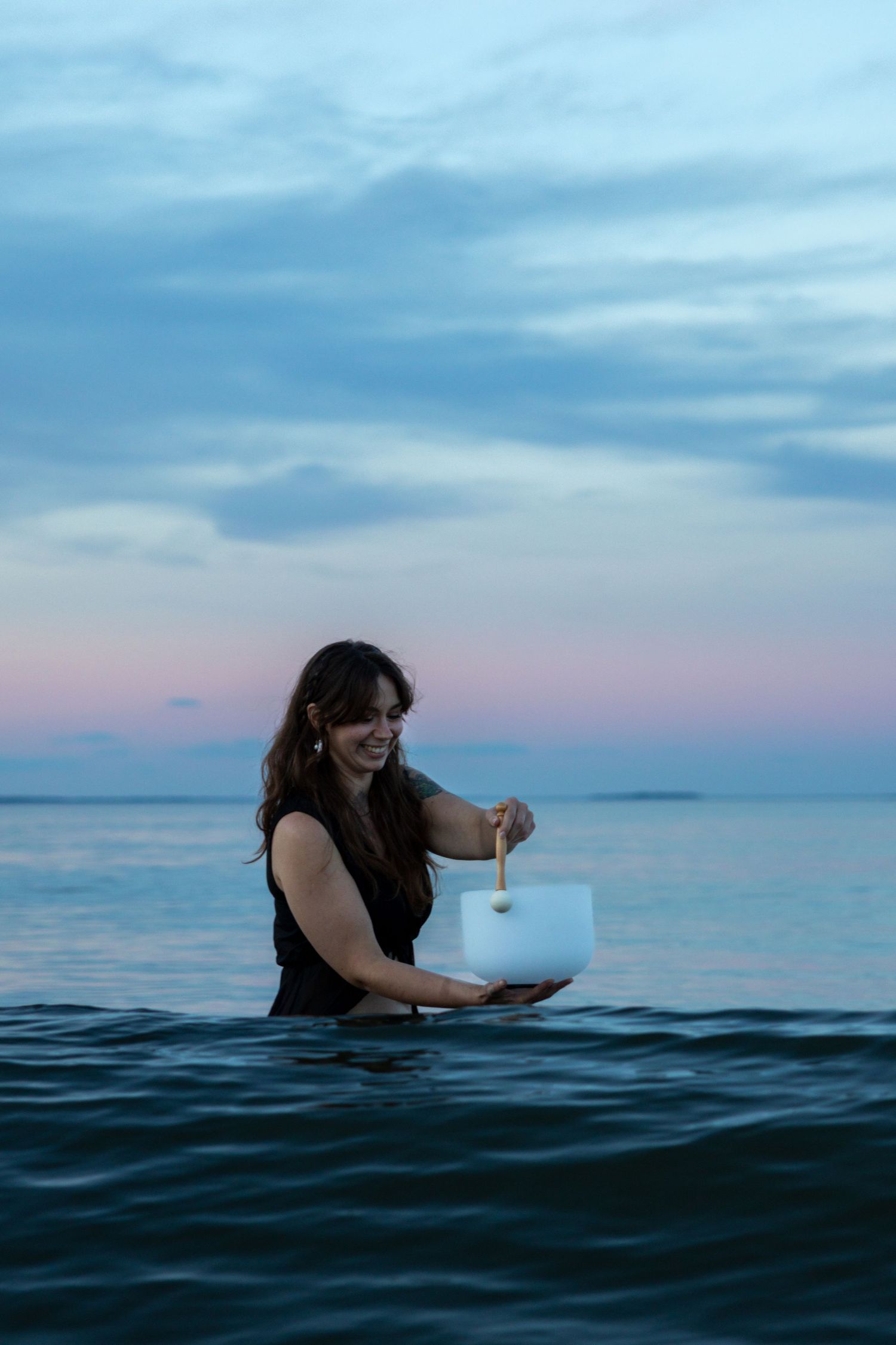 Woman standing waist-deep in ocean at dusk, smiling and holding a white handbag, with a pastel cloudy sky backdrop.
