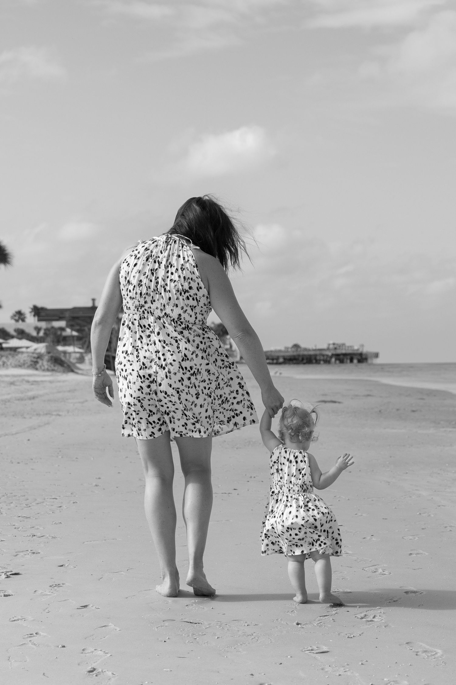 Black and white photo of a mother and daughter in matching dresses walking on a beach.