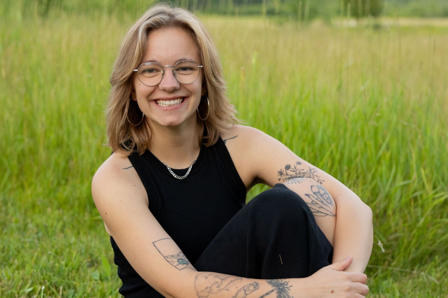 Smiling woman with glasses and tattoos sits cross-legged in a lush green meadow wearing black.
