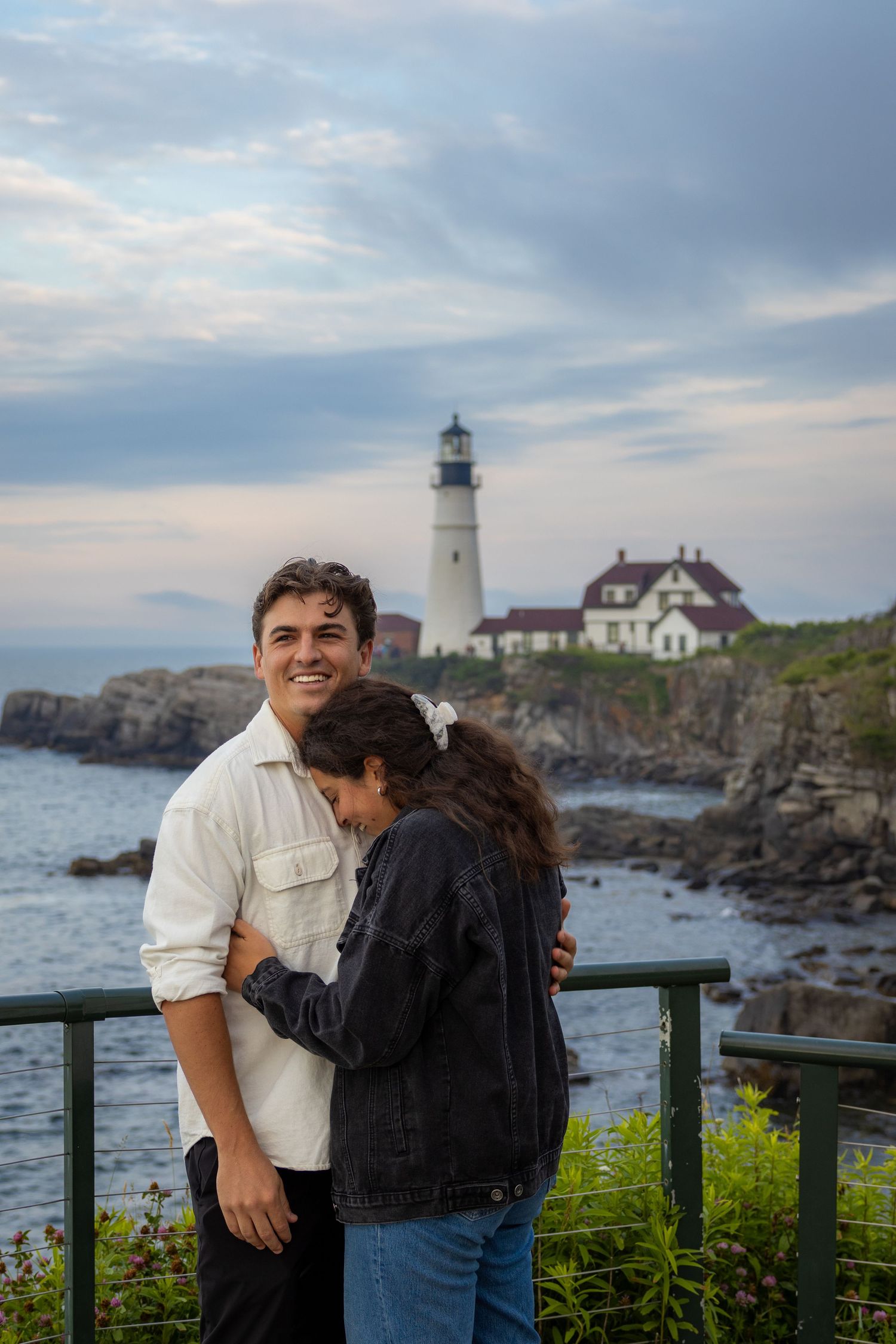 Couple embracing near a railing with Portland Head lighthouse and rocky coastline in the background at sunset.