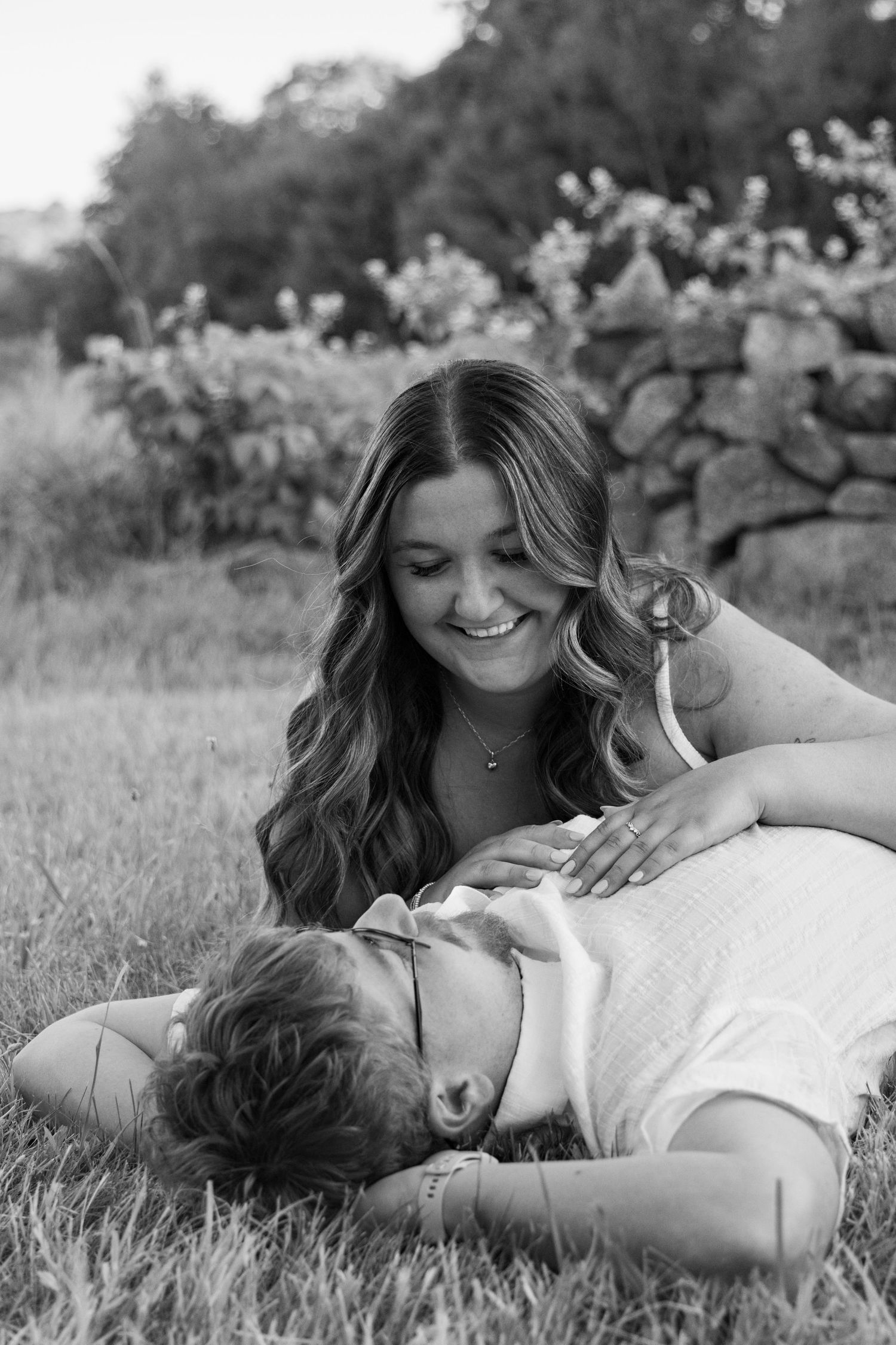 Black and white photo of a woman smiling while lying on a man resting in the grass outdoors.