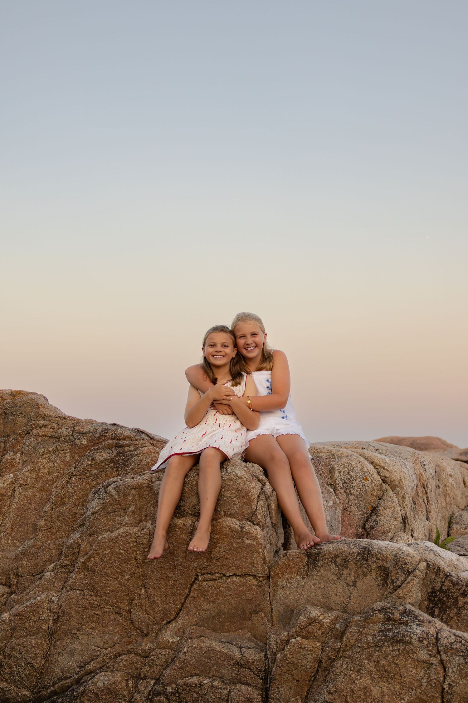 Two girls in white dresses sitting on rocks hugging and smiling at sunset.