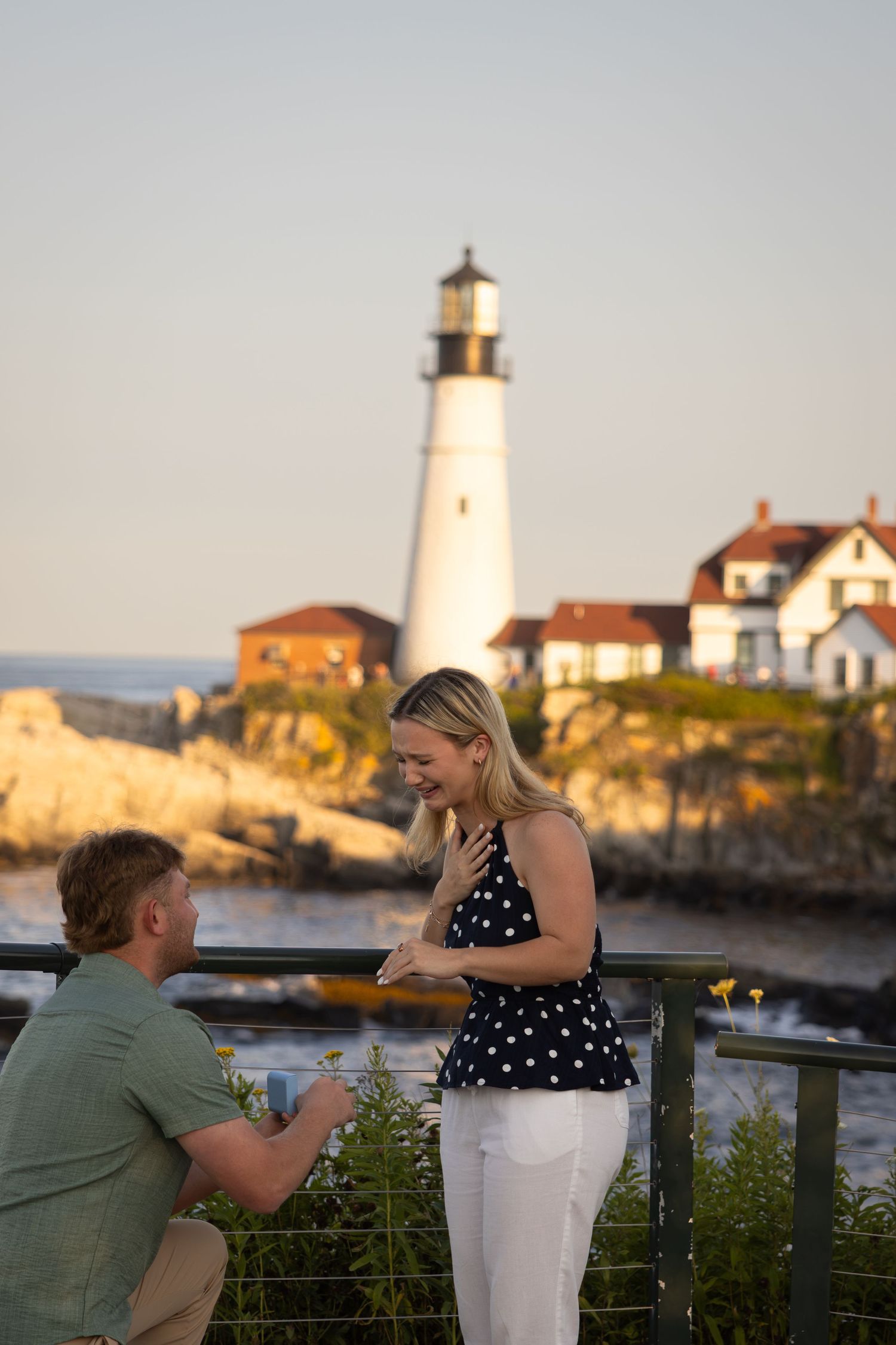 Man proposes to woman on bended knee at Portland Head Light lighthouse in Maine at golden hour.