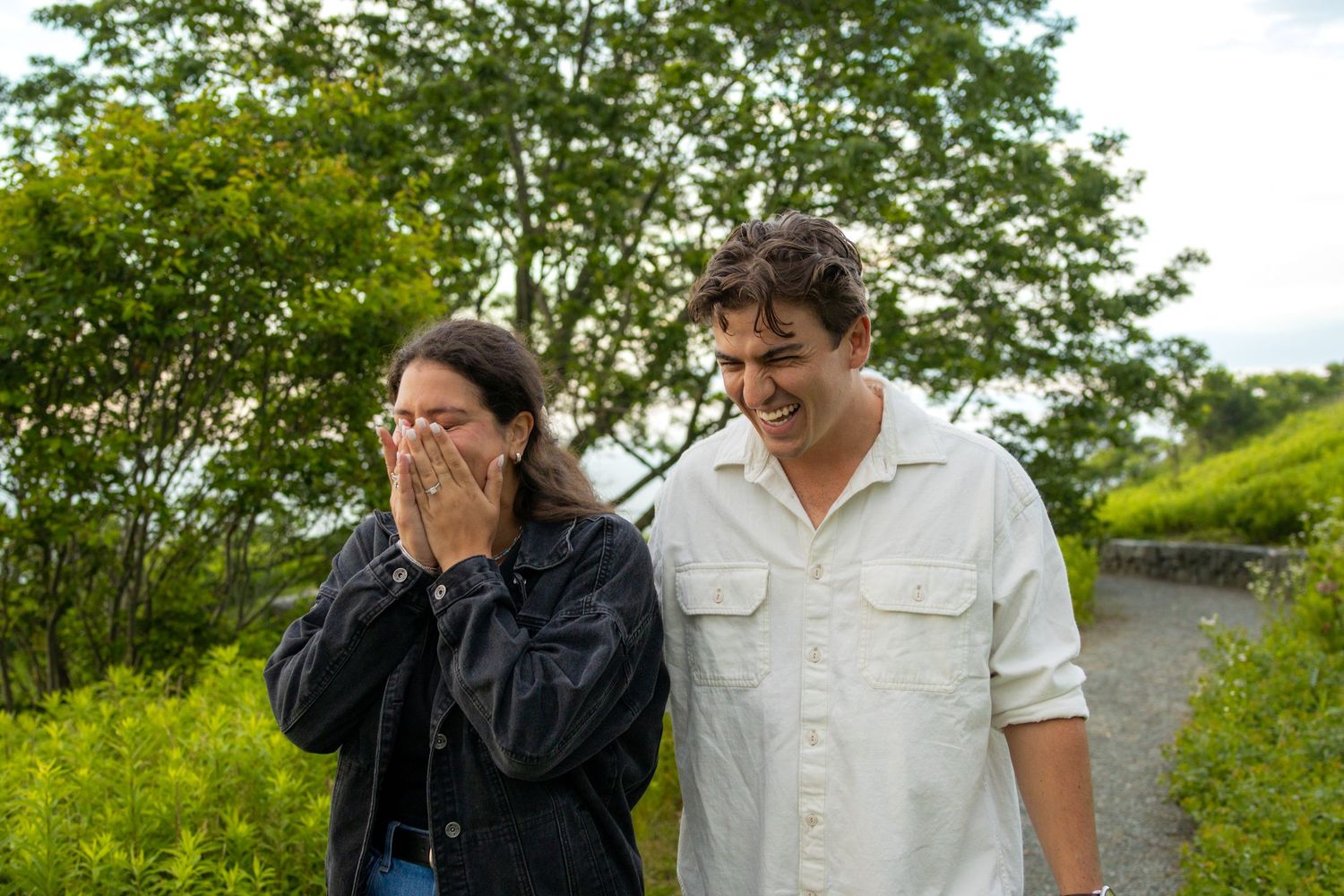 Couple laughs together on a green tree-lined path, woman covering her face with her hands in joy.