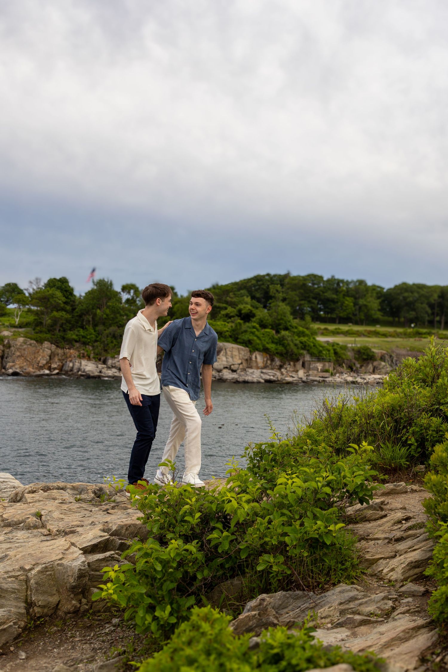 Two young men walking and talking along a rocky coastal path with lush greenery and calm water nearby.