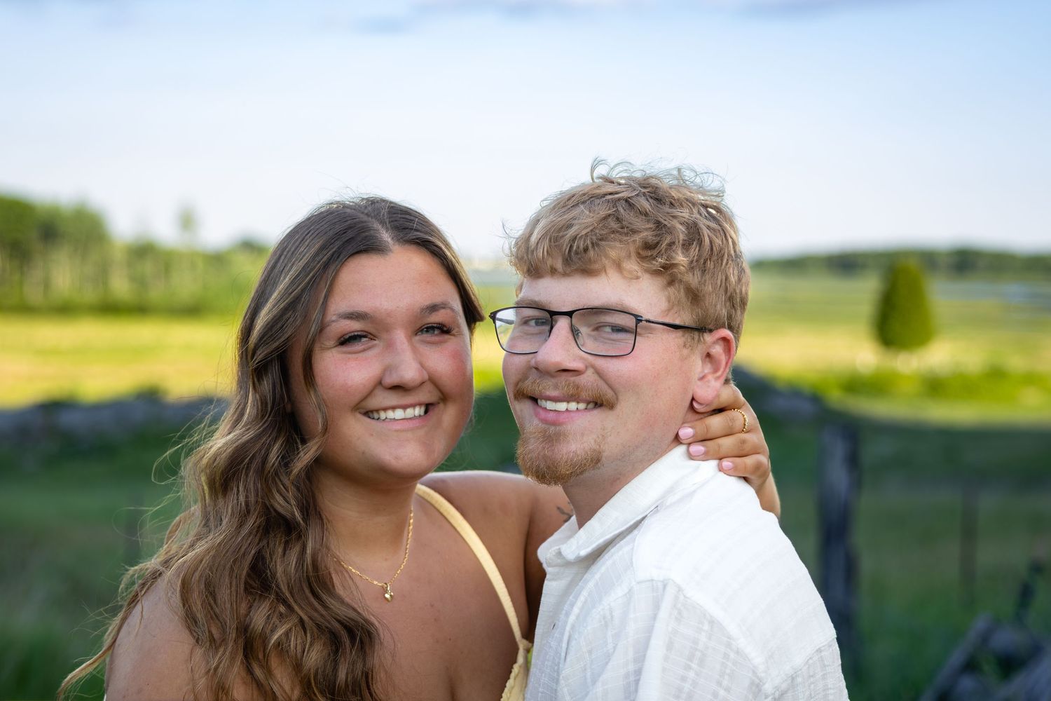 A smiling young couple poses outdoors near a green field on a sunny day, the woman in yellow and man in white shirt.