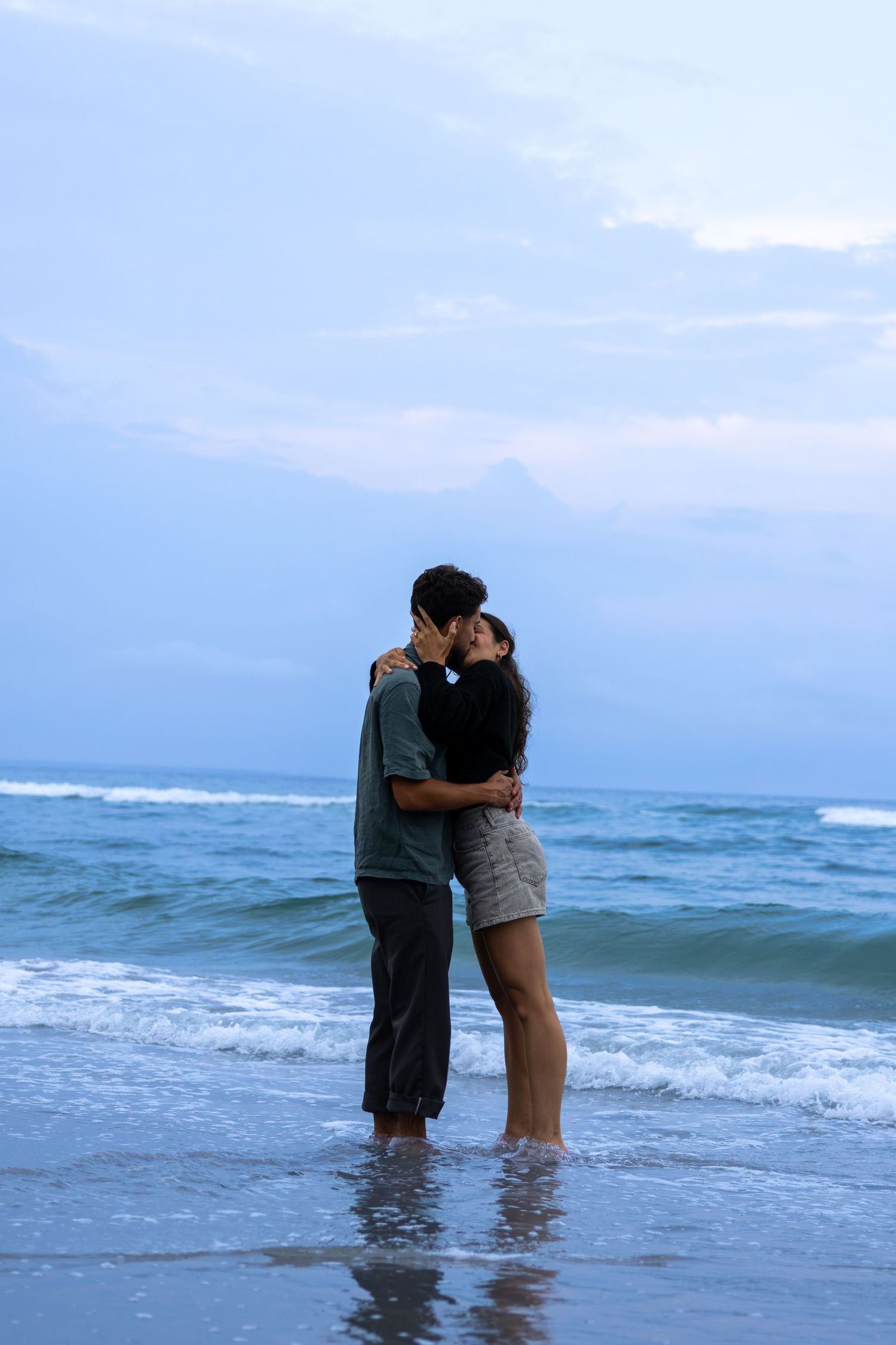 A romantic couple kissing and embracing on a sandy beach at dusk with gentle ocean waves in the background.