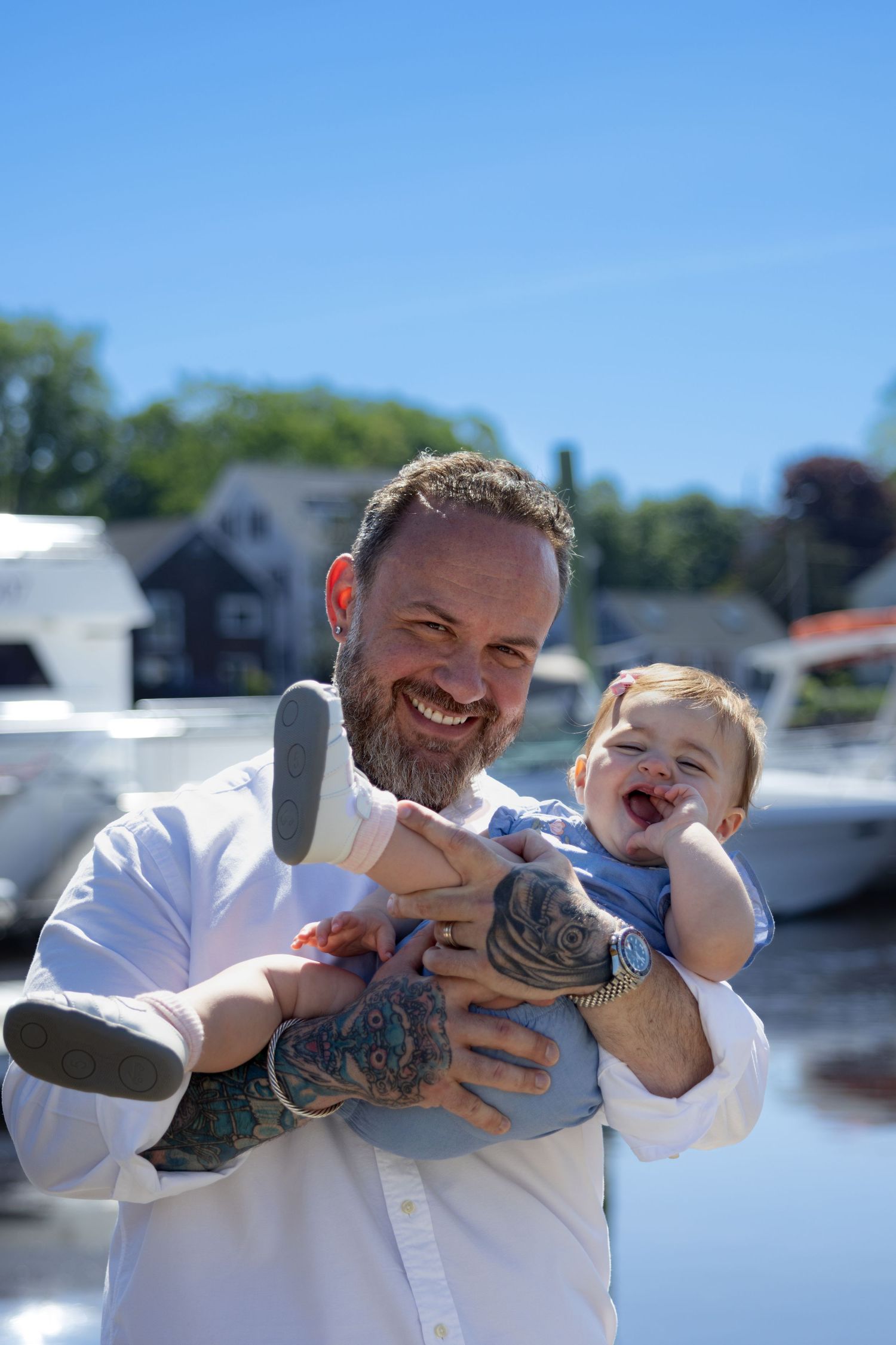 Smiling bearded tattooed man holding a laughing baby outdoors near a marina on a sunny day.