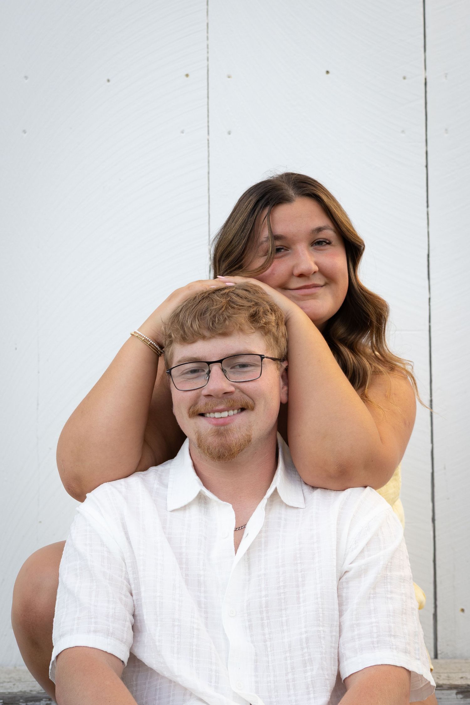 Woman playfully holds mans head from behind as he smiles at the camera against a white wooden wall.