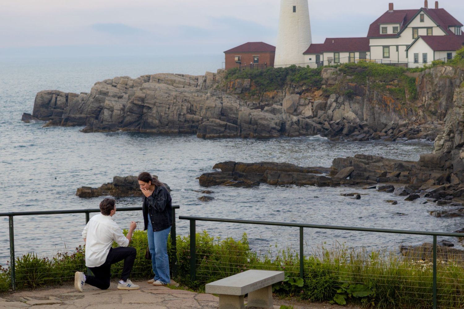 Man proposes on one knee to woman at a scenic coastal lighthouse overlook at dusk.