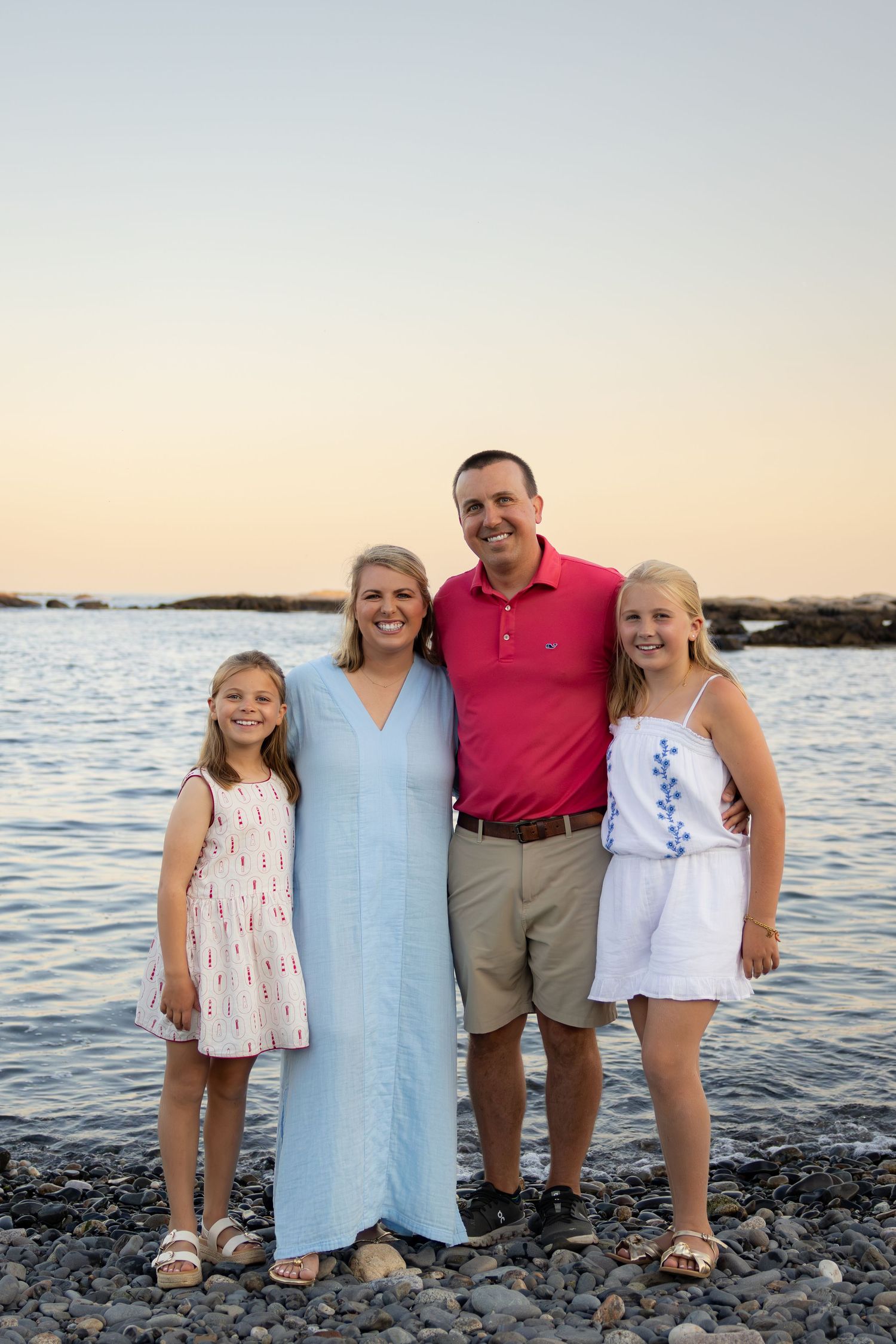Family of four smiling together on a rocky beach at sunset, wearing summer outfits in white, blue, and pink.