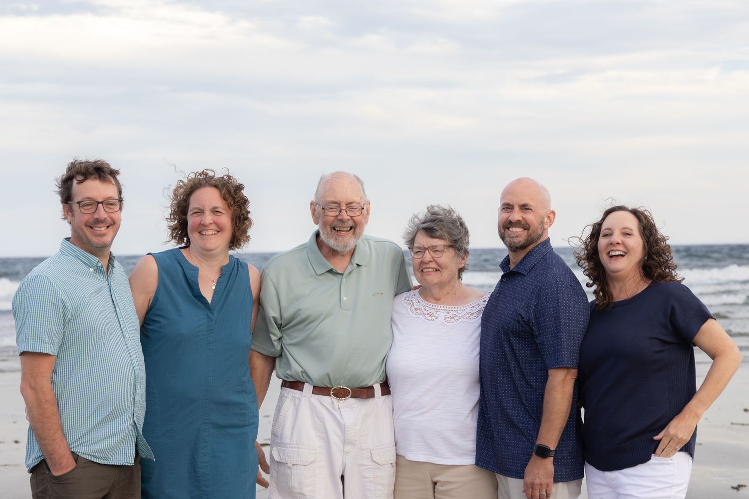 Six smiling family members pose together on a sandy beach with ocean waves and cloudy sky in the background.