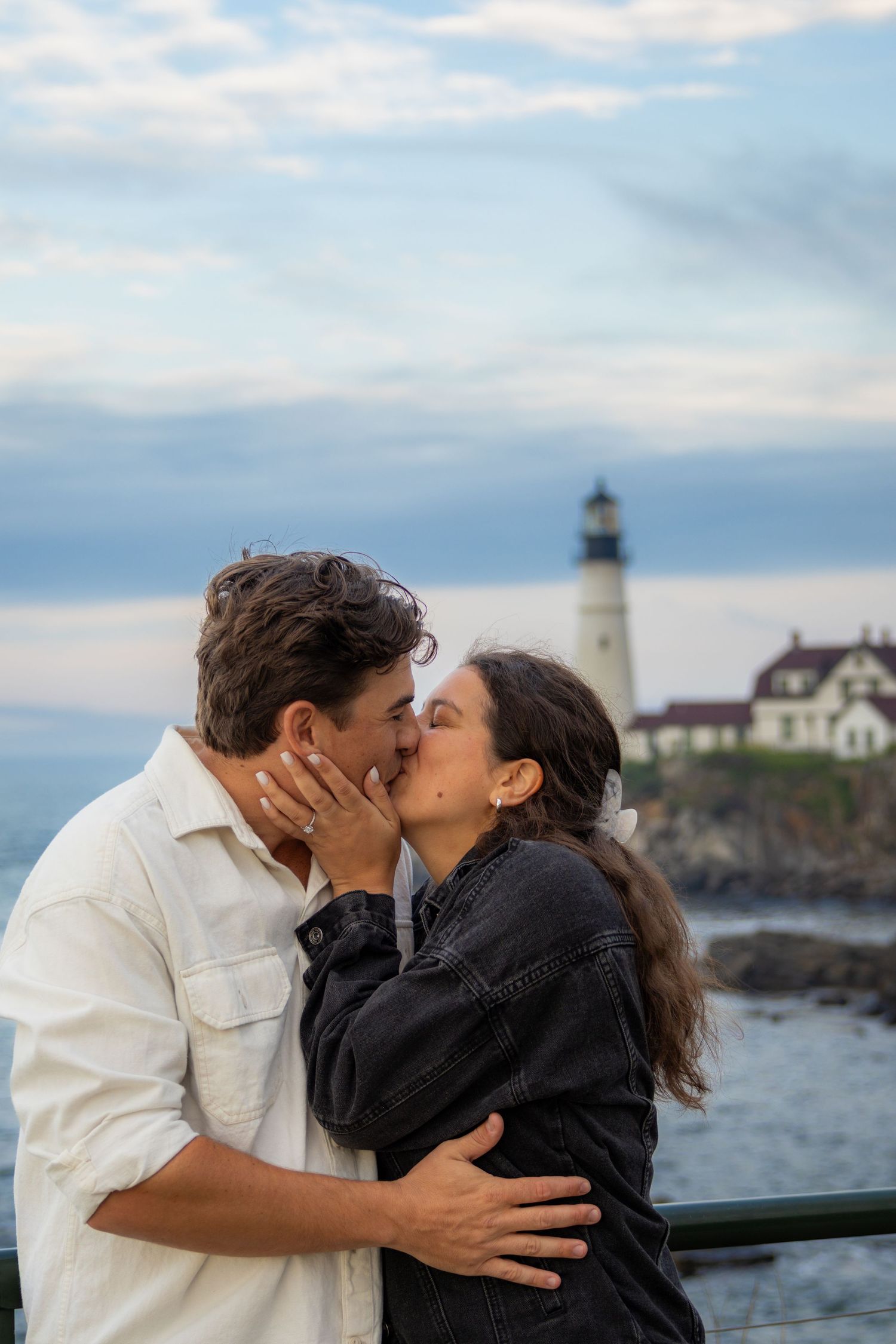 Couple sharing a romantic kiss in front of a classic white lighthouse by the ocean at dusk.