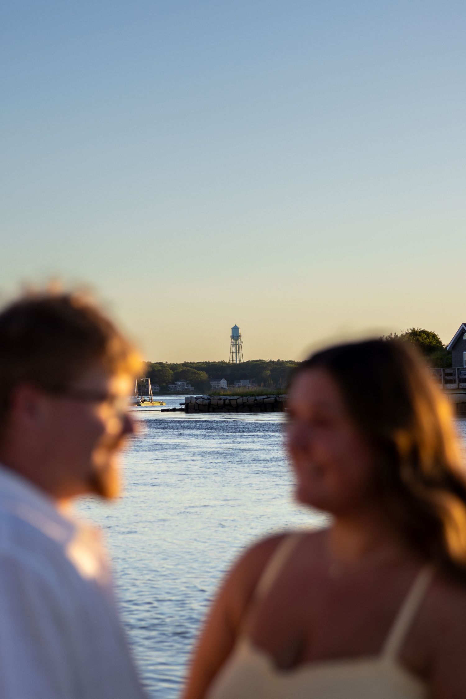 Couple stands out of focus in the foreground with a lighthouse and calm harbor visible at sunset.