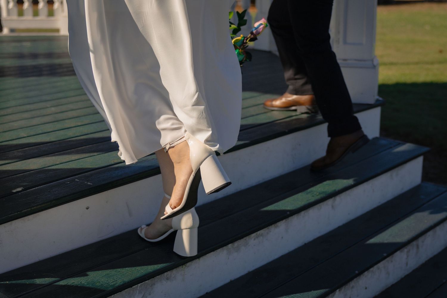 Close-up of a brides white heeled shoes and grooms brown shoes walking up sunlit outdoor steps.