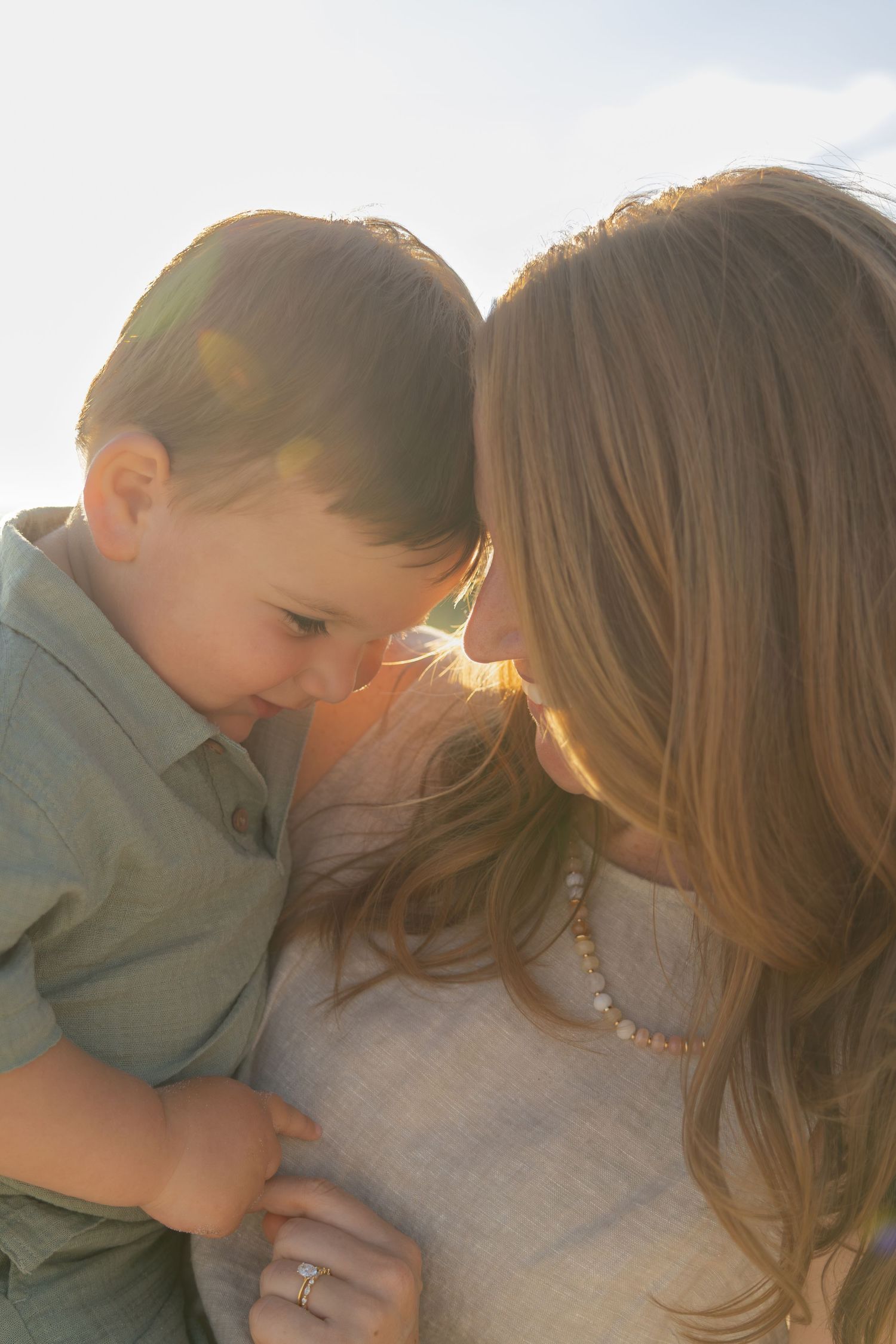 A mother and young son share a tender forehead-to-forehead moment in warm golden backlit sunlight outdoors.