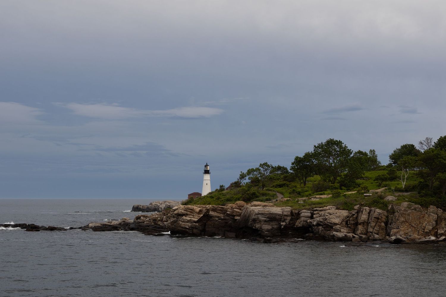 A white lighthouse stands on a rocky coastal cliff surrounded by trees under a cloudy sky.