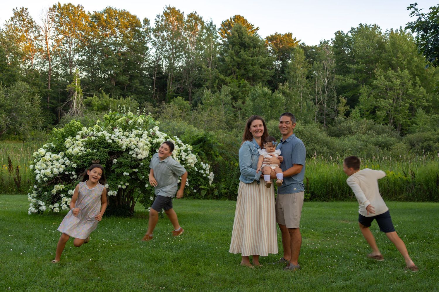 Family of five posing outdoors on lush green lawn with blooming white hydrangeas and trees in background at dusk.