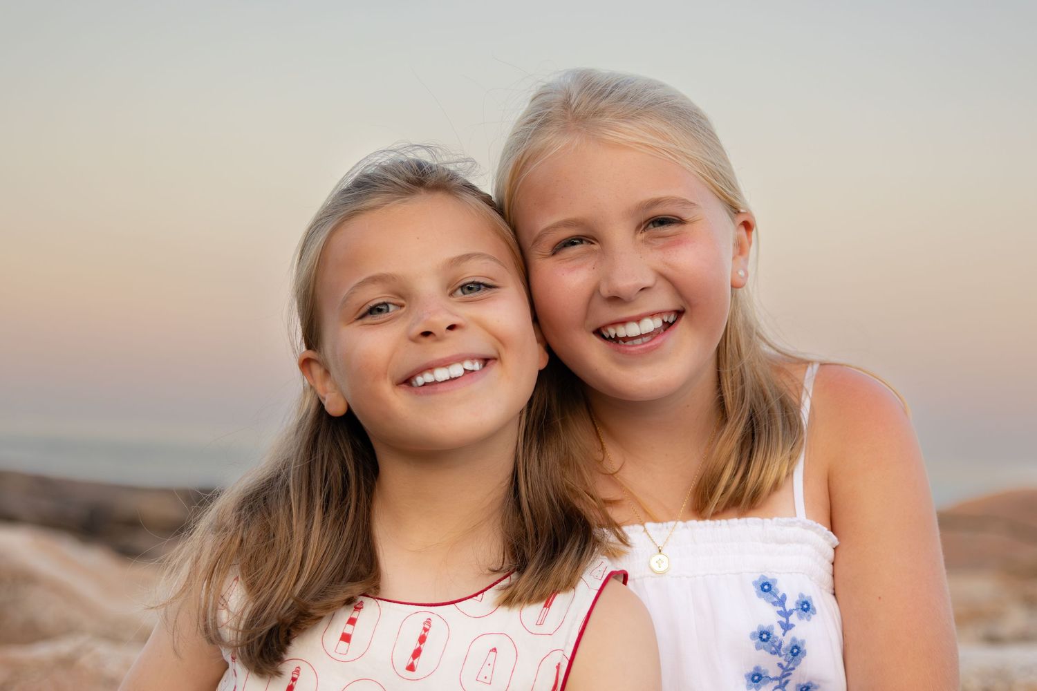 Two smiling blonde girls in white dresses pose together on a sandy beach at sunset.