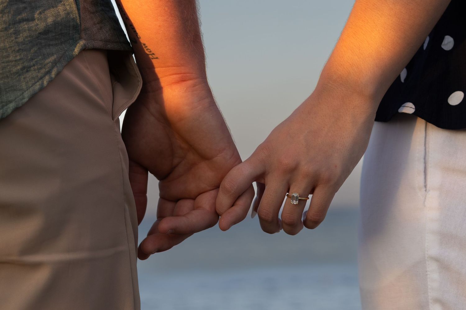 Couple holding hands with engagement ring visible, romantic beach sunset in background.