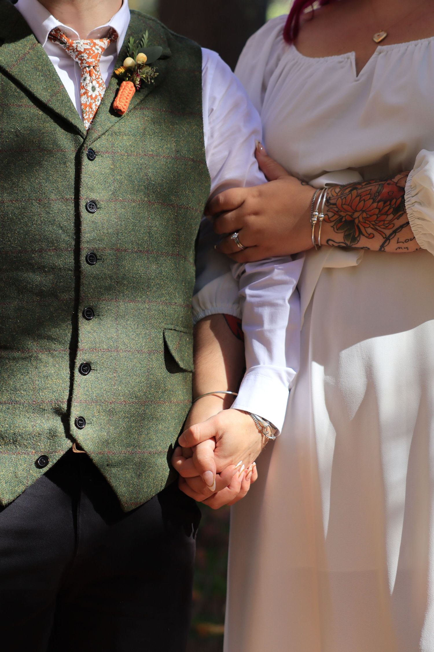 Couple holds hands closely, groom in green tweed vest with floral tie, bride in white dress with tattoo.