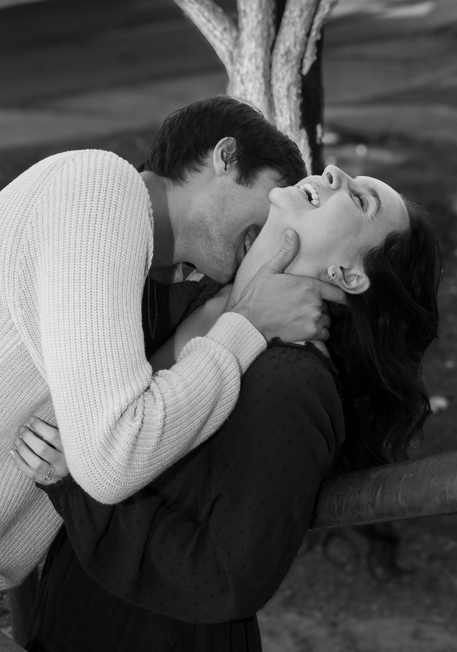 Black and white romantic photo of a man kissing a womans neck as she laughs and leans back outdoors.