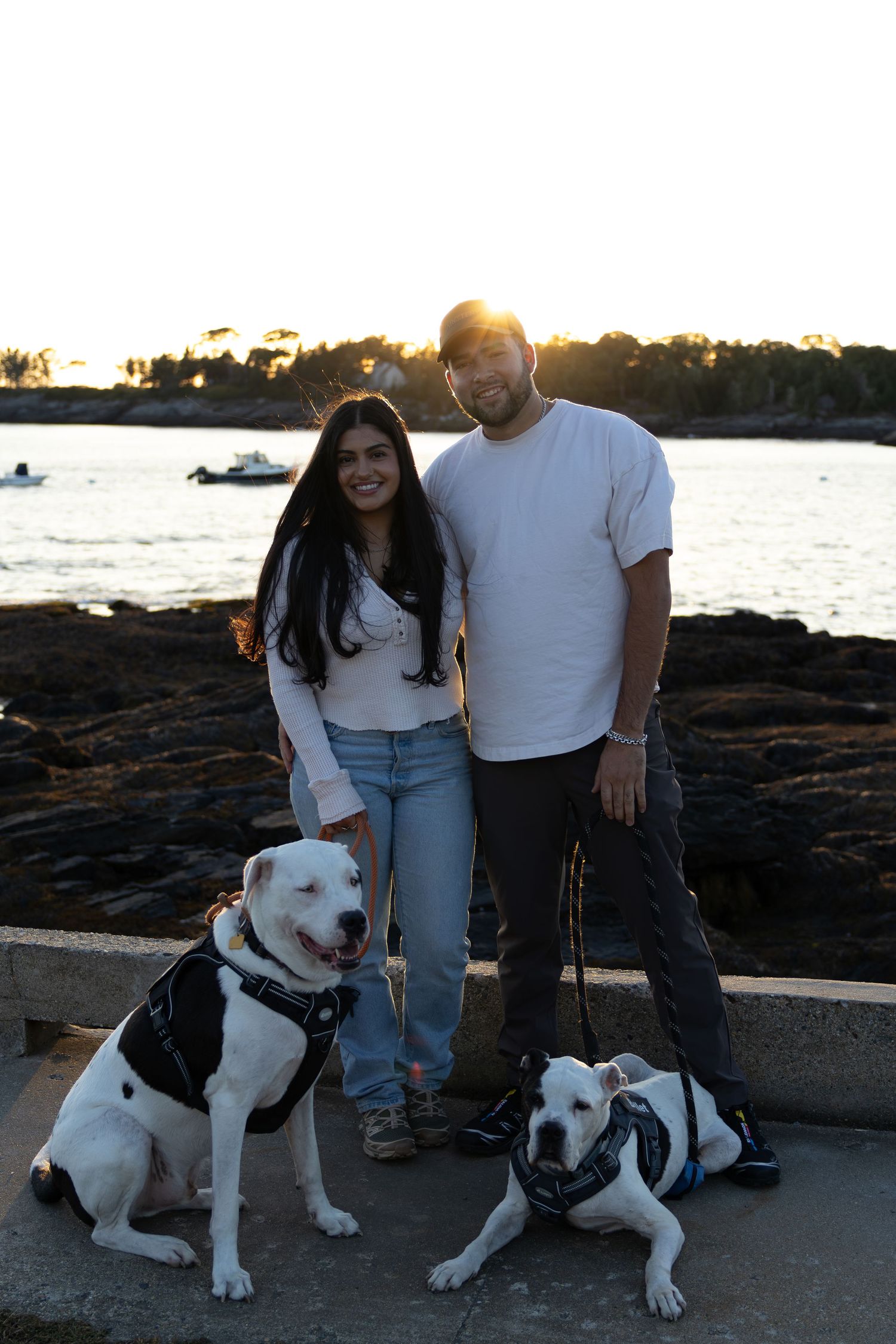 Couple smiling with two white pit bulls on leashes by rocky waterfront at sunset.