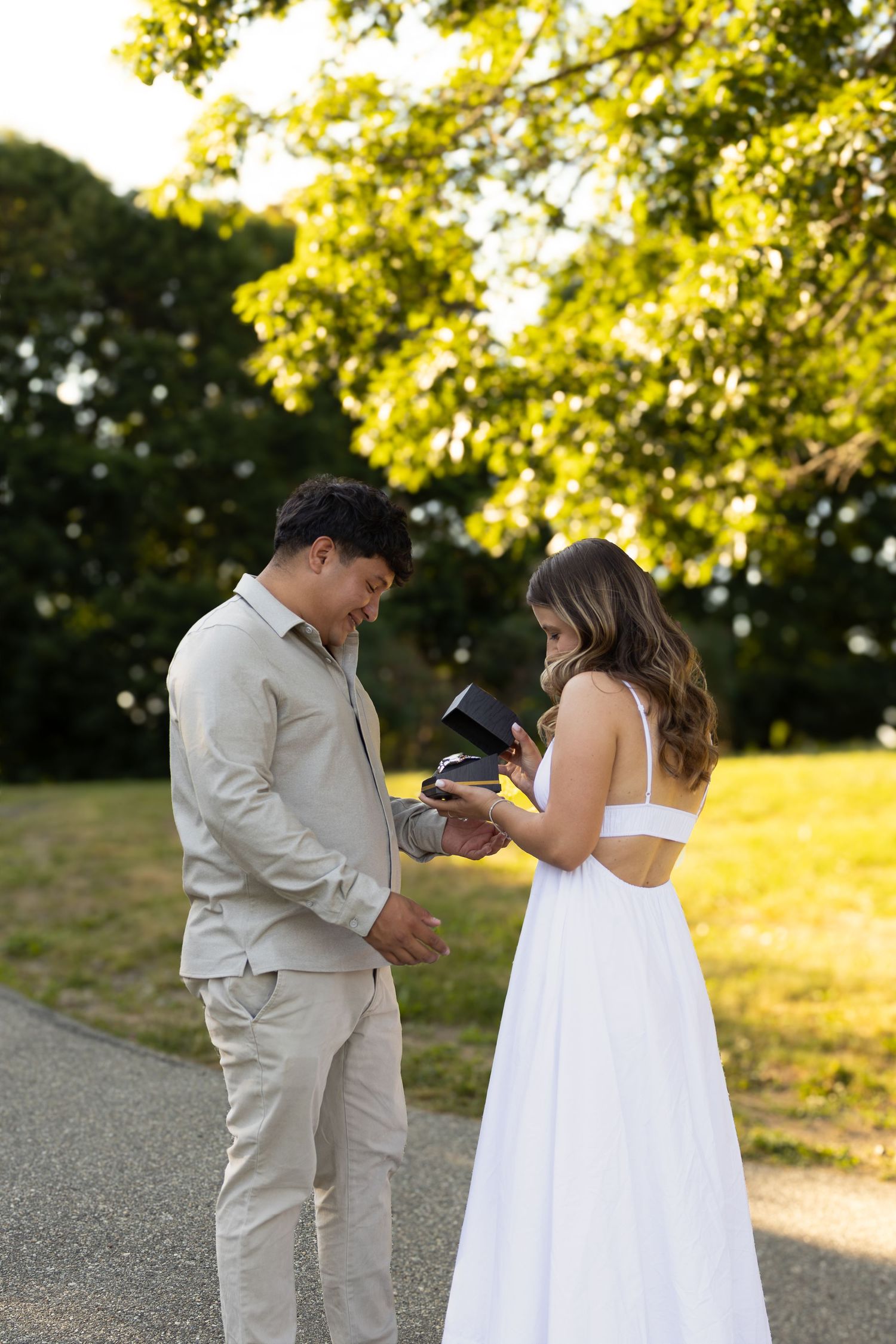 Couple in formal wear exchanging rings outdoors, man in beige suit and woman in white dress, surrounded by lush greenery.