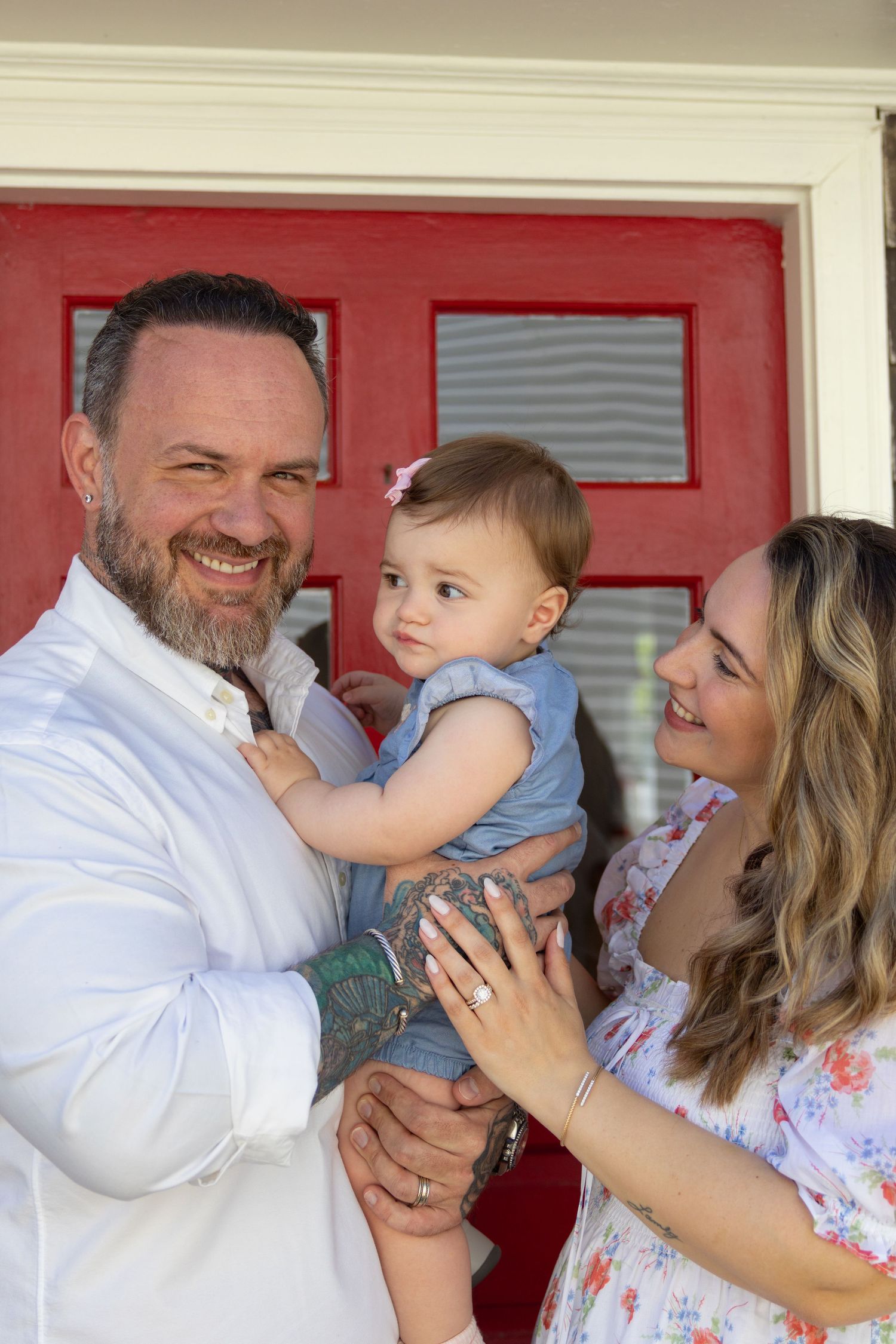 Smiling couple in front of a red door holding their baby girl in a denim outfit.