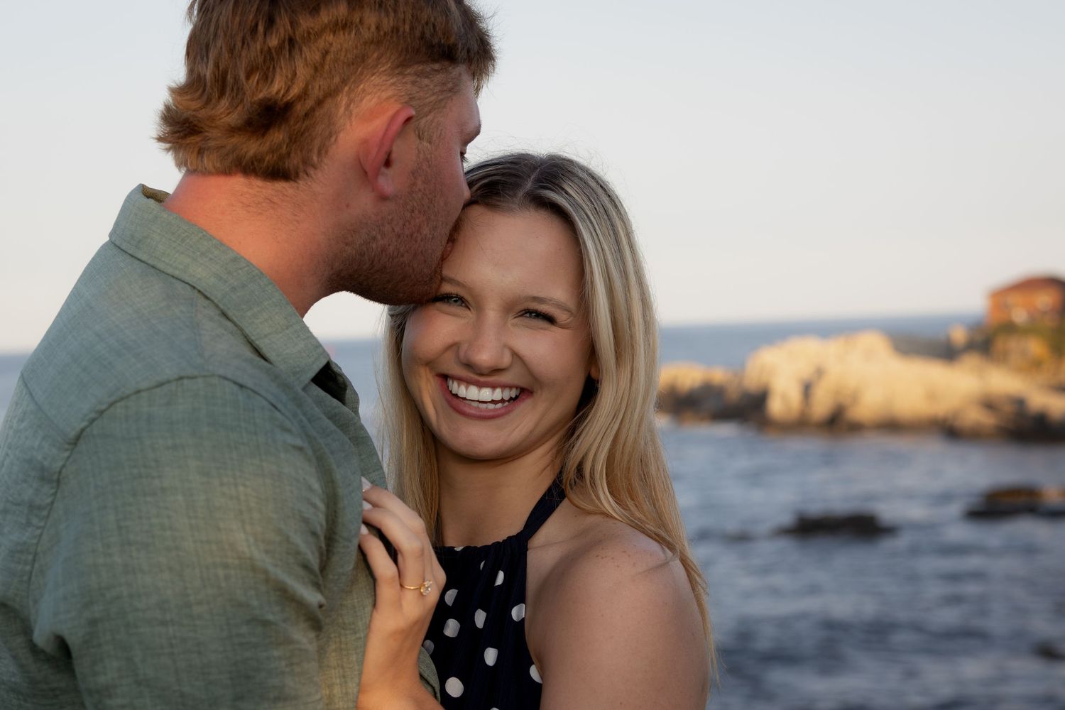 Couple sharing a romantic moment by the ocean at sunset, woman smiling joyfully as man kisses her cheek.