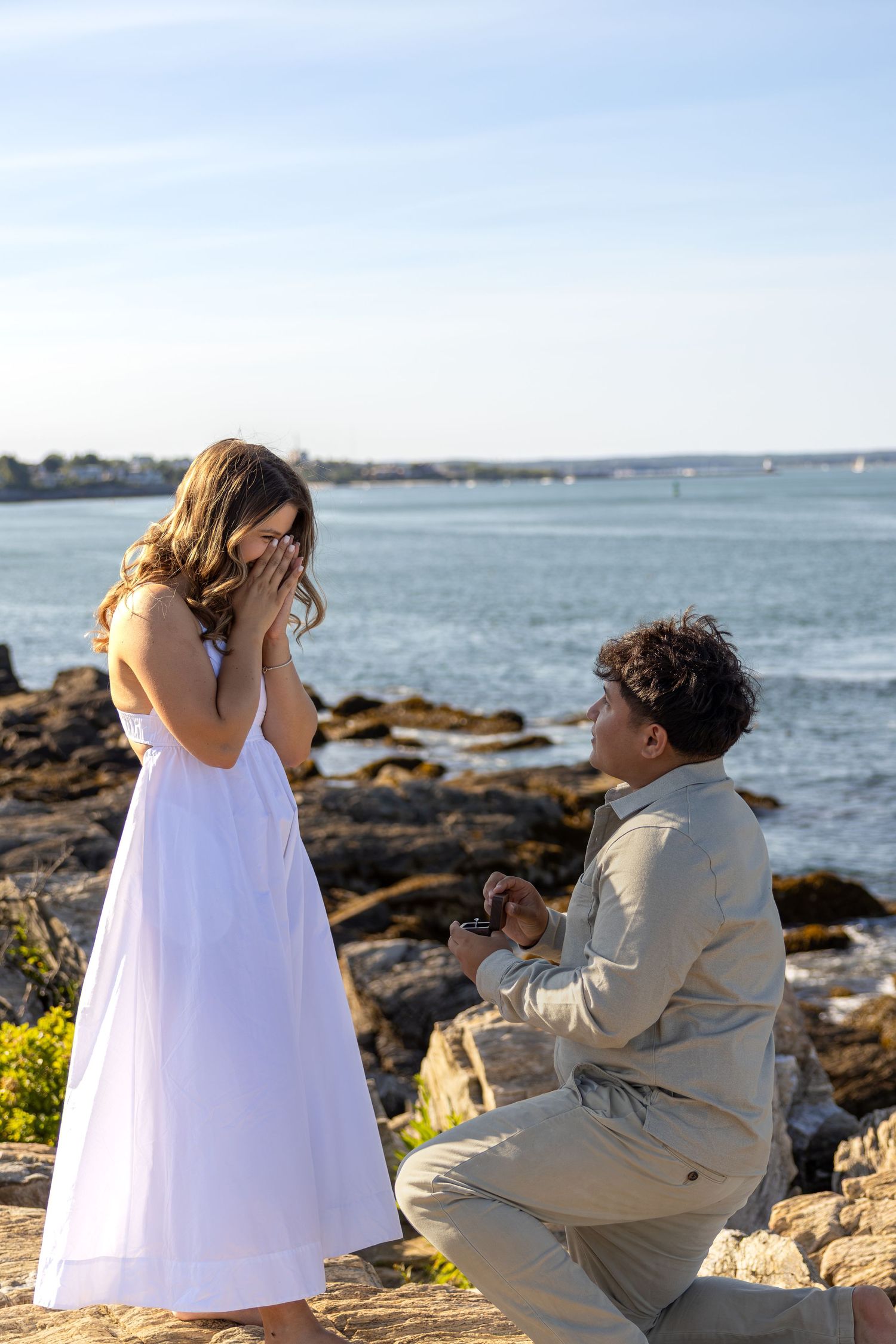 Man kneeling on rocky shore proposing to emotional woman in white dress with ocean backdrop.