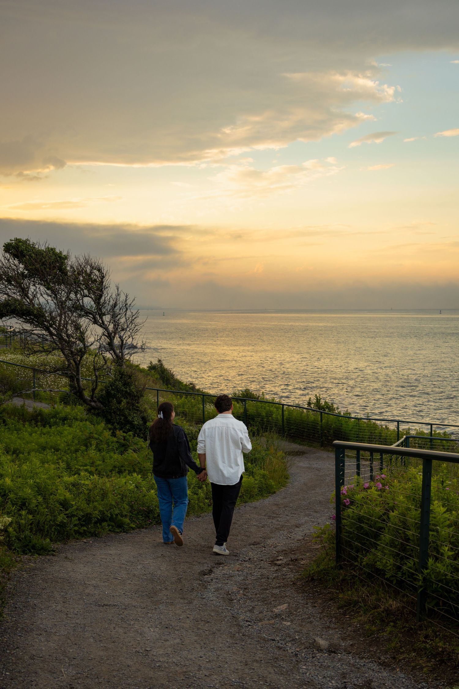 Couple walking hand in hand along a coastal path toward a stunning golden sunset over the ocean.