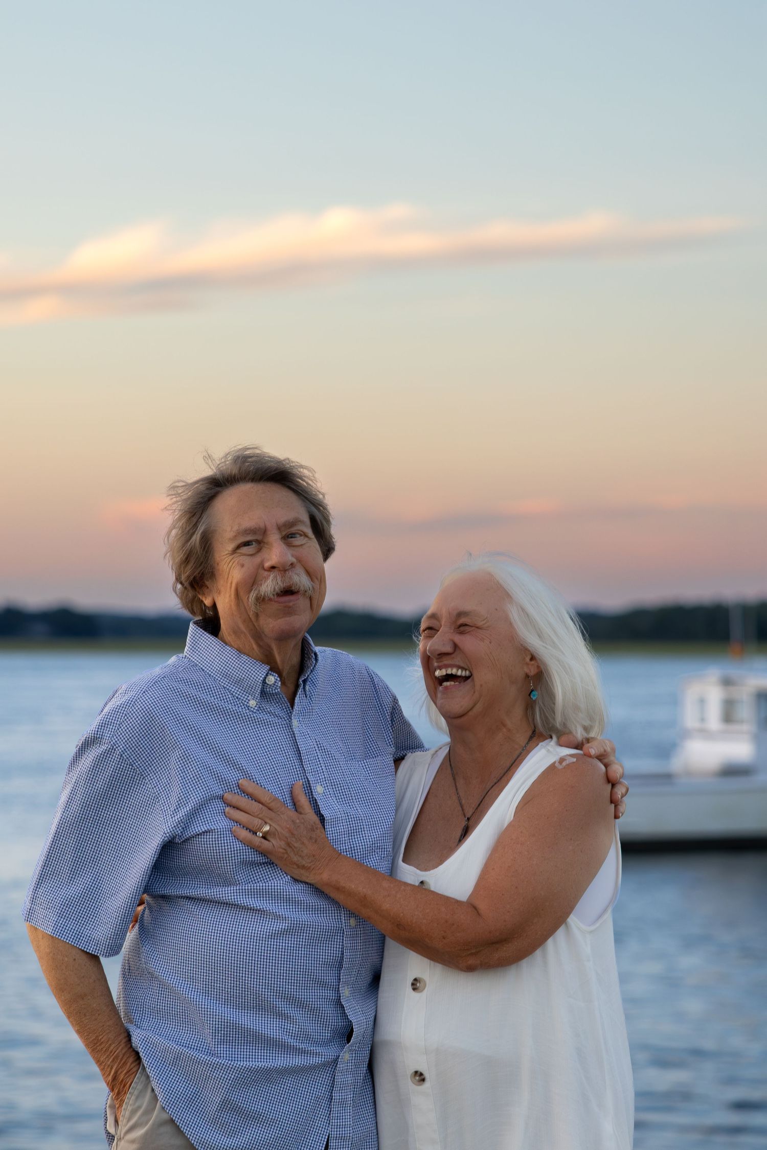 Happy senior couple embracing and laughing together by a waterfront at sunset, enjoying a romantic evening outdoors.