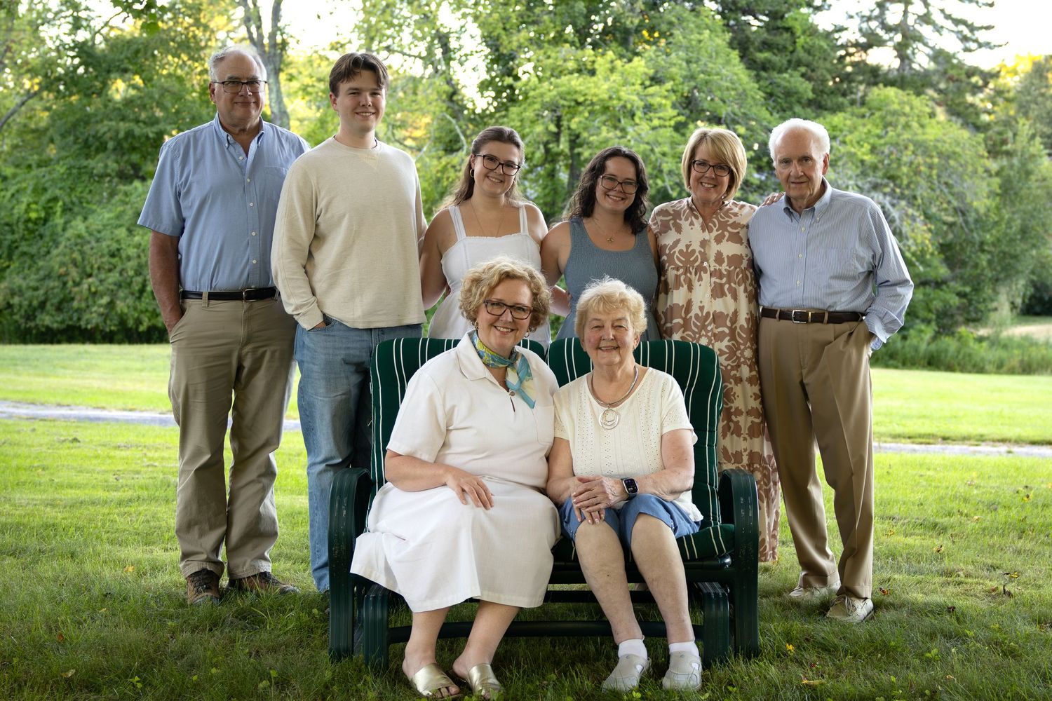 Multi-generational family group portrait outdoors on green lawn with trees, two elderly women seated on bench, others standing behind.