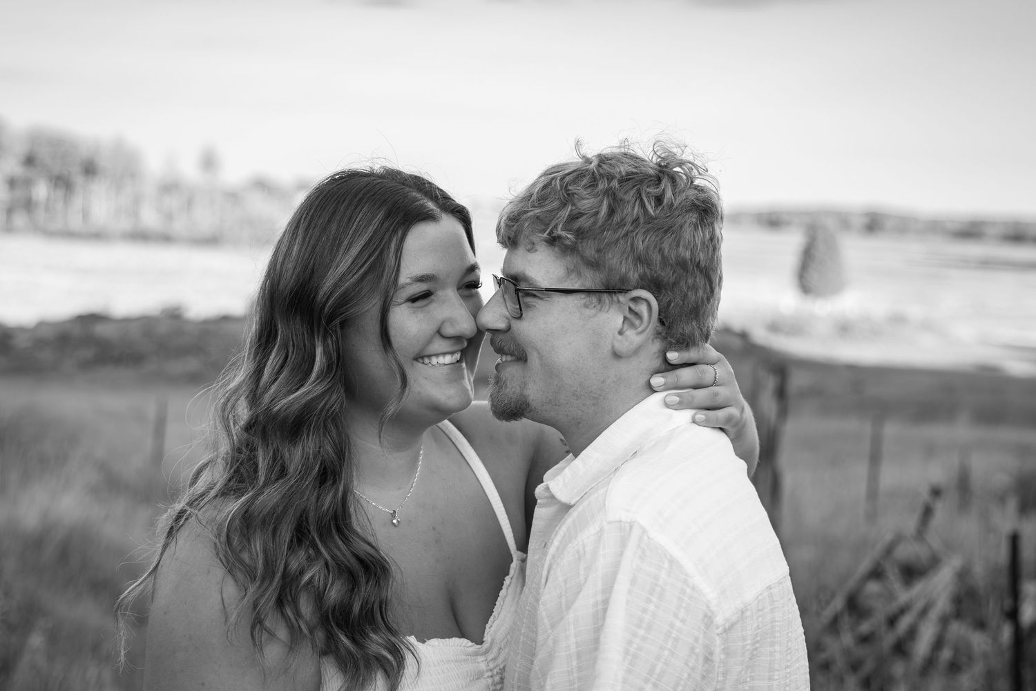 Black and white romantic photo of couple smiling and embracing near a coastal beach landscape.