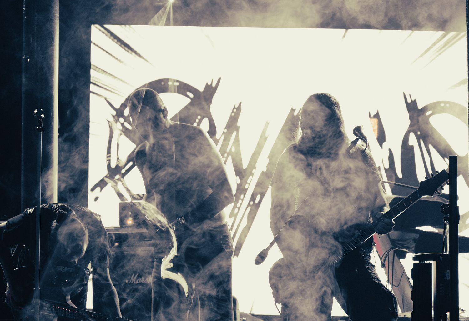 Two performers shrouded in smoke on dramatic backlit stage with industrial metal props.