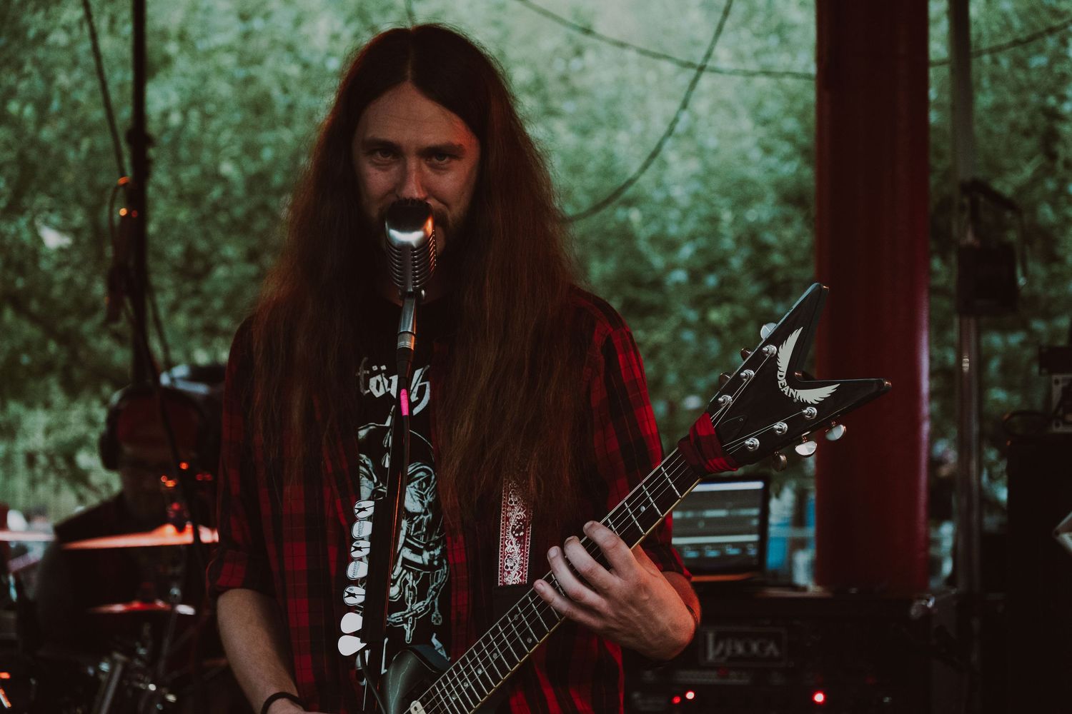 Long-haired male guitarist in red flannel shirt playing black electric guitar at outdoor concert.