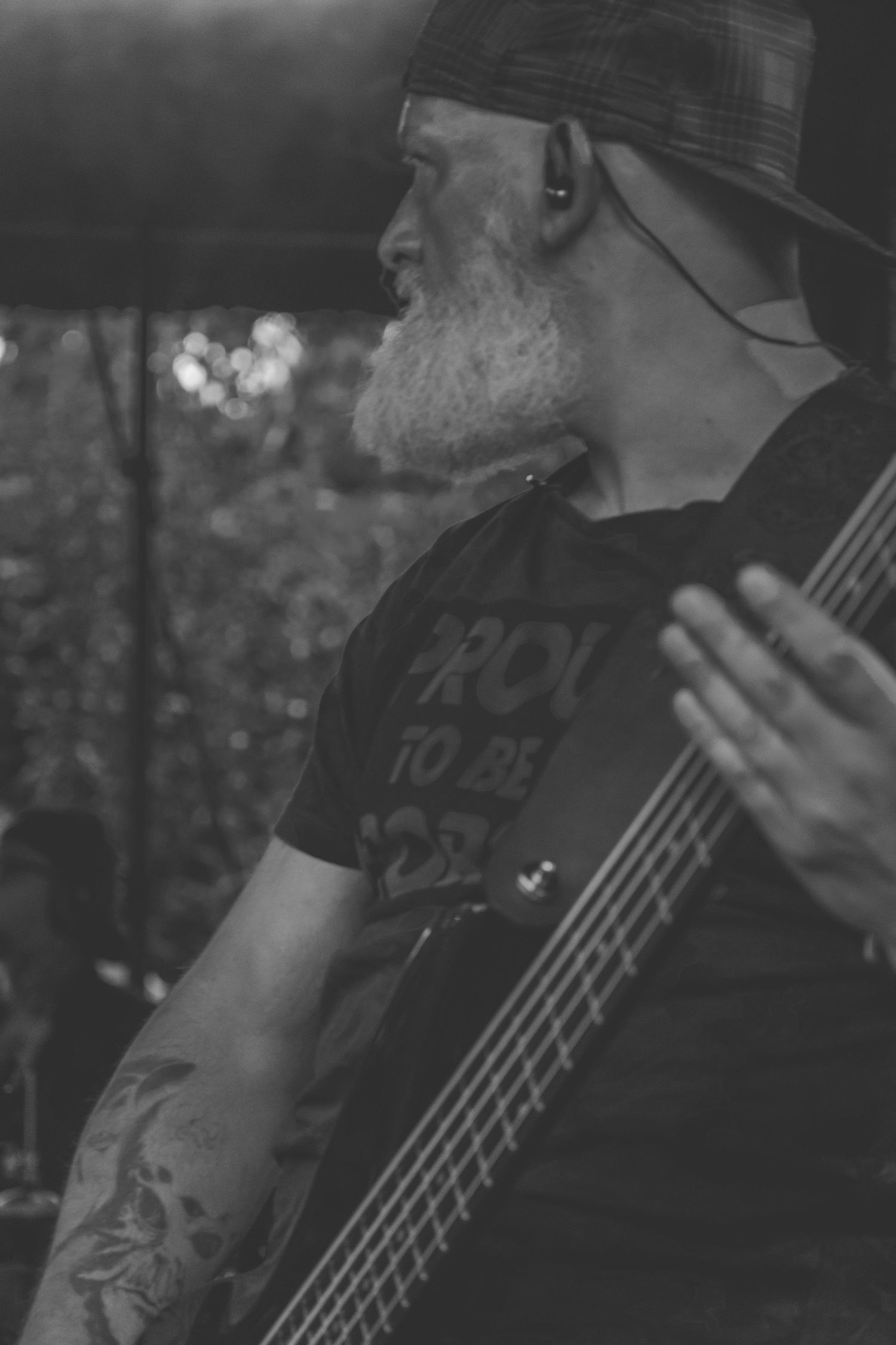 Black and white photo of bearded bassist with tattoos playing bass guitar on stage.