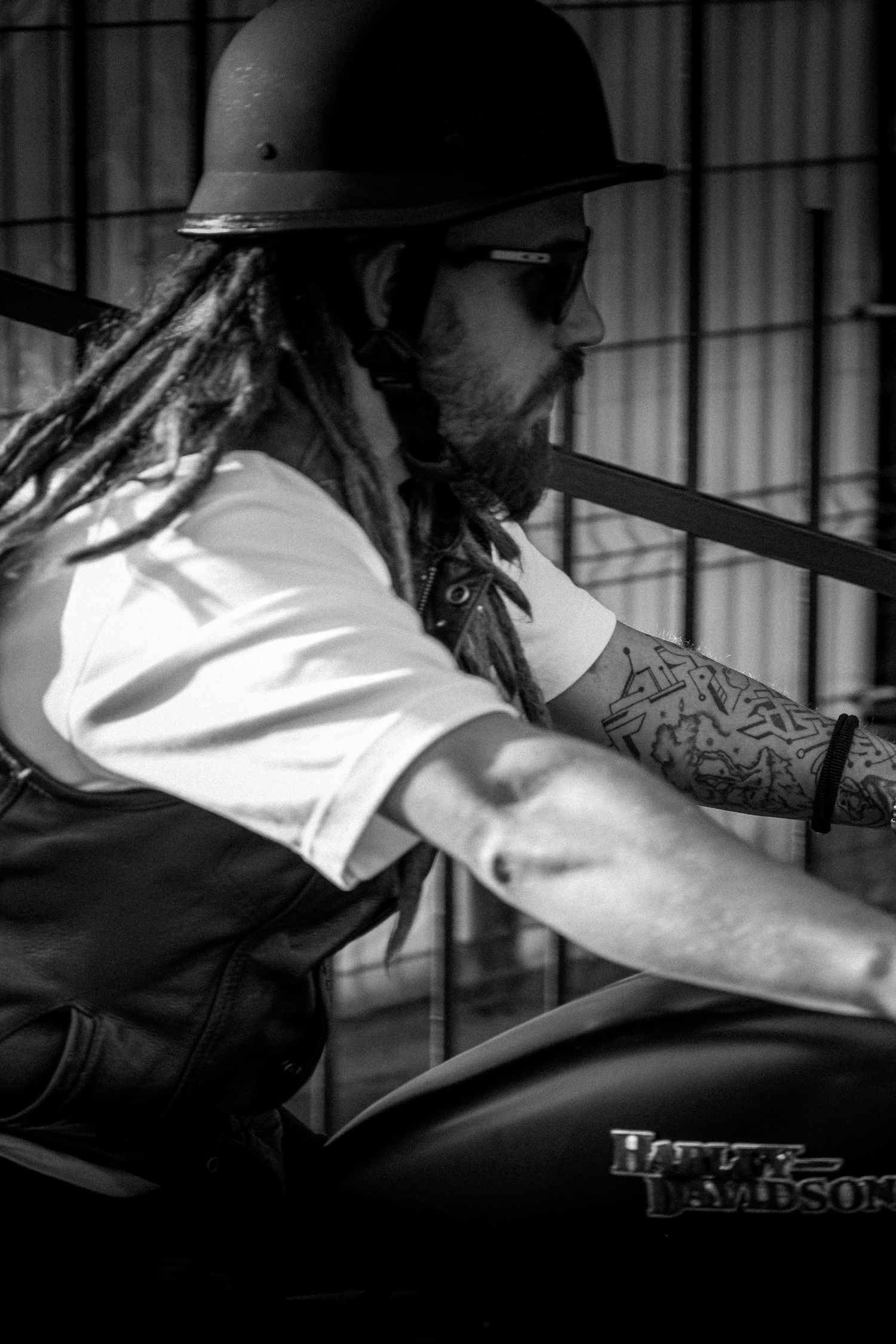 Tattooed biker with dreadlocks and helmet rides a Harley-Davidson motorcycle in black and white photo.