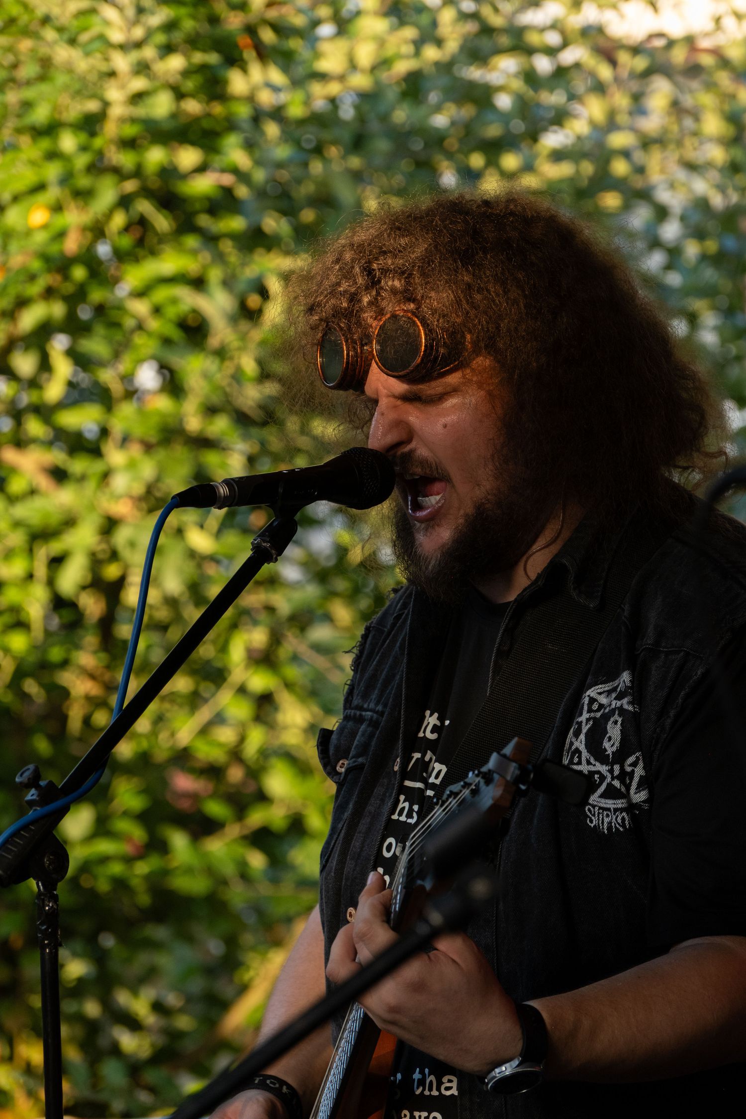 Curly-haired male guitarist with round steampunk goggles singing into microphone at outdoor concert.