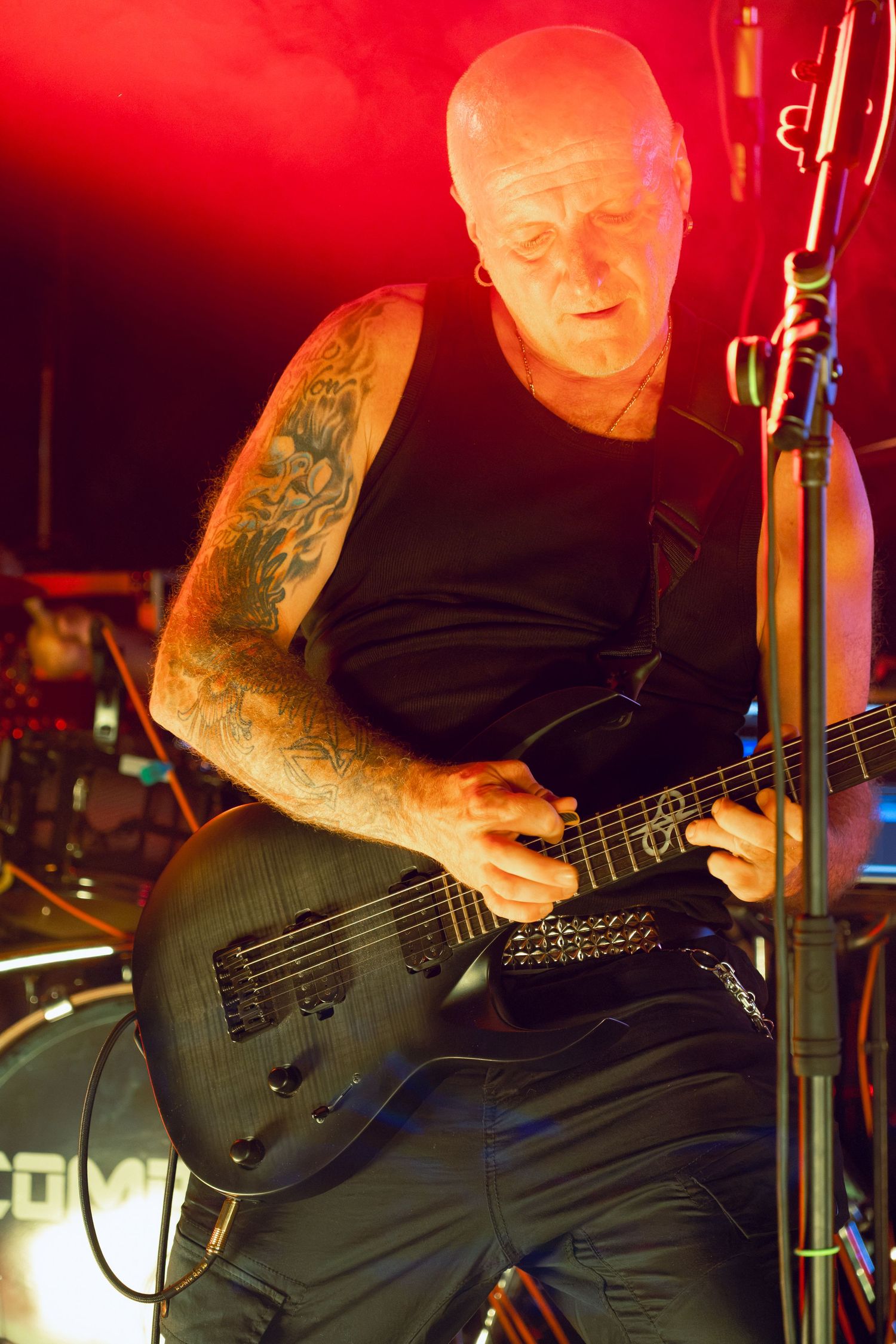 Tattooed bald male guitarist playing black electric guitar under red stage lights at concert.