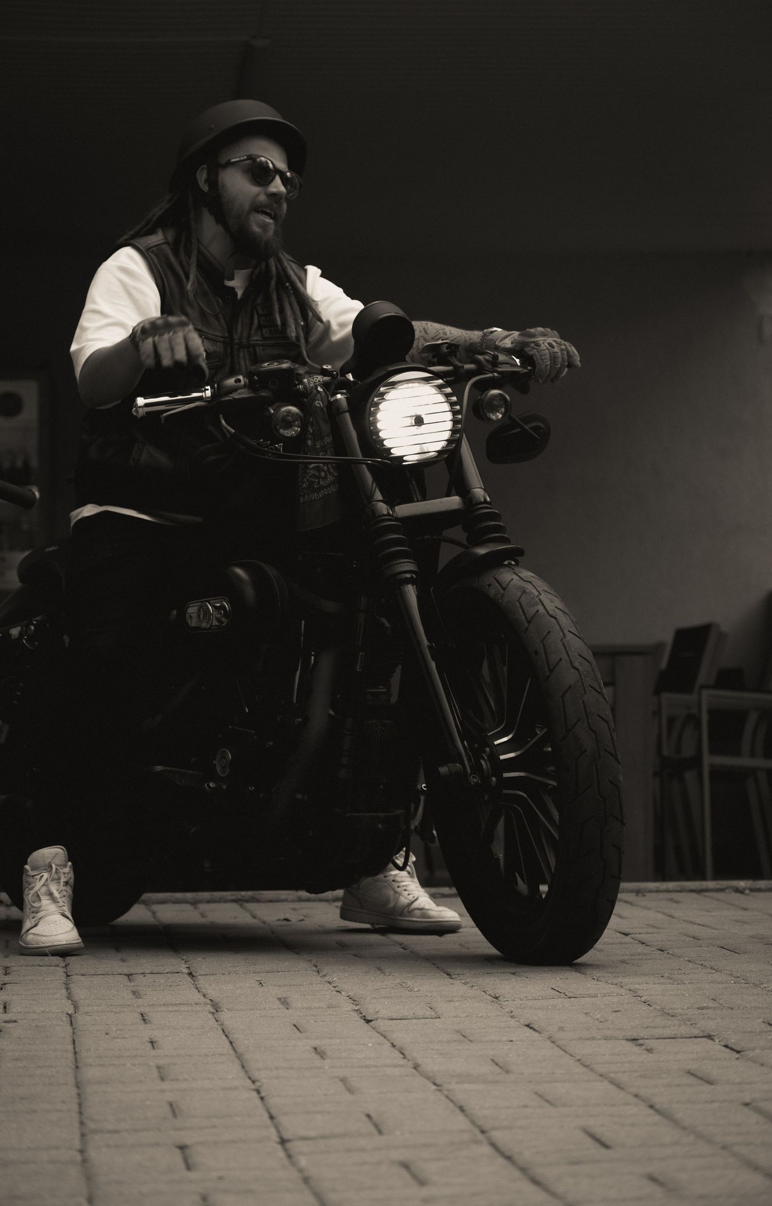 Black and white portrait of dreadlocked man on black custom motorcycle with headlight on.