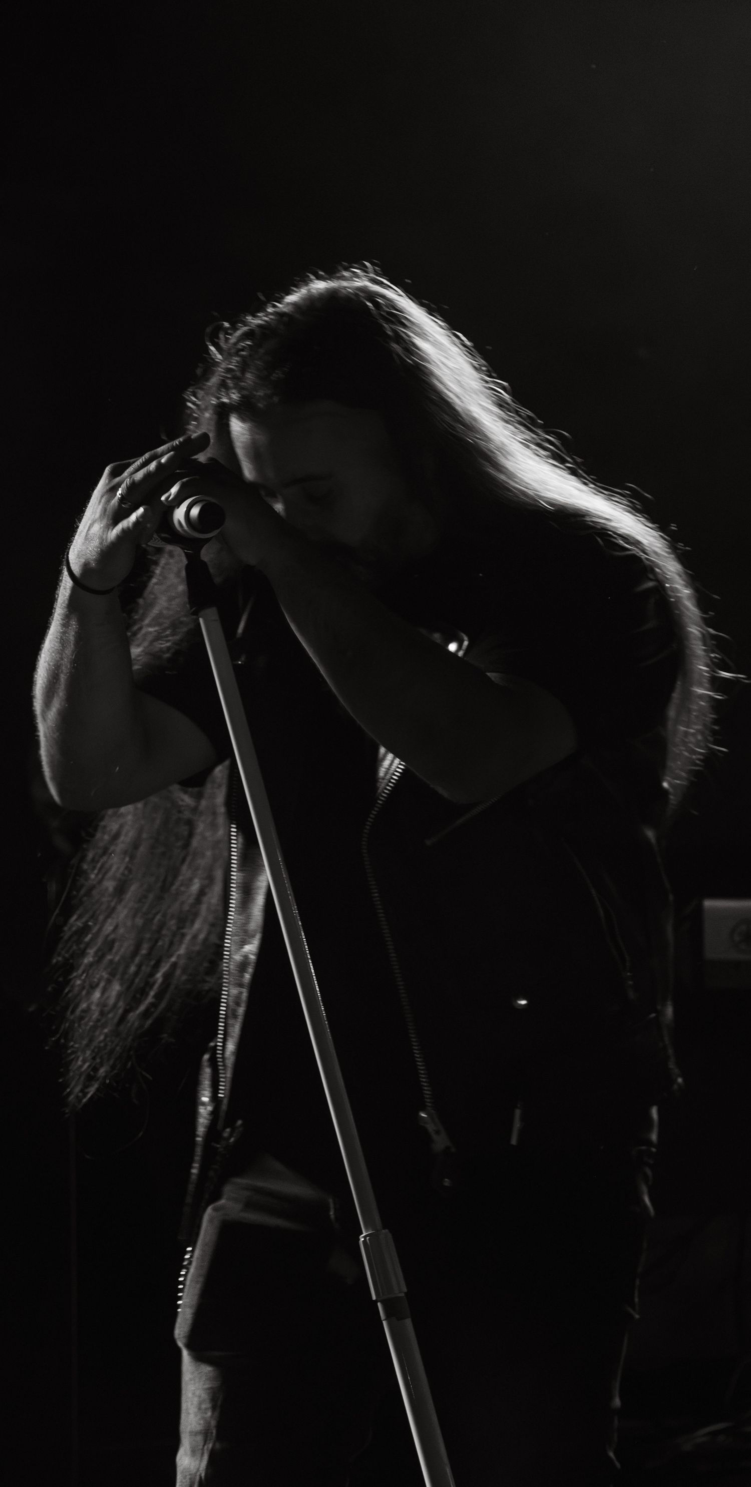 Black and white photo of a long-haired metal musician bowing at a microphone stand.