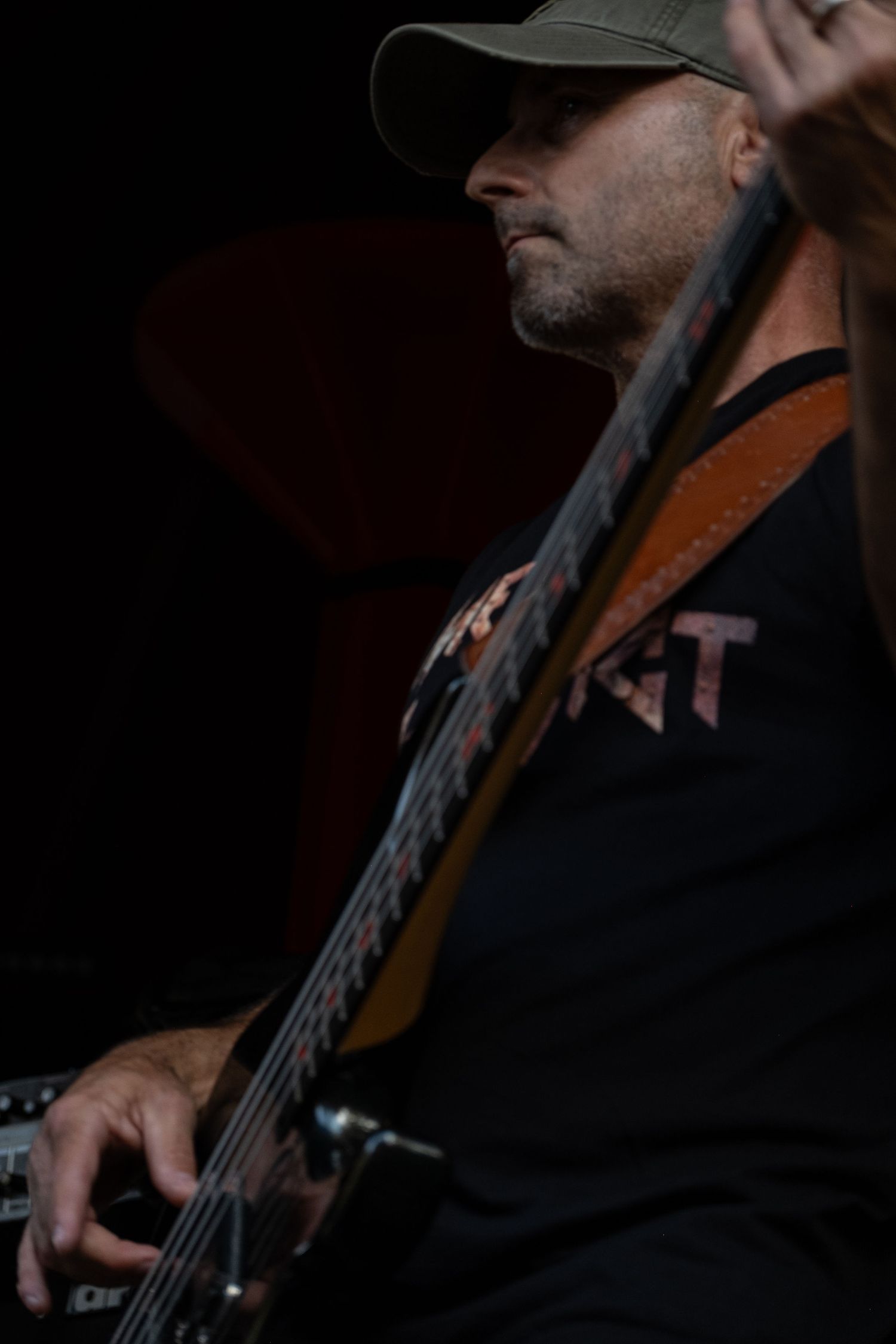 Male bassist wearing a cap performs on stage in low light at a live music concert.
