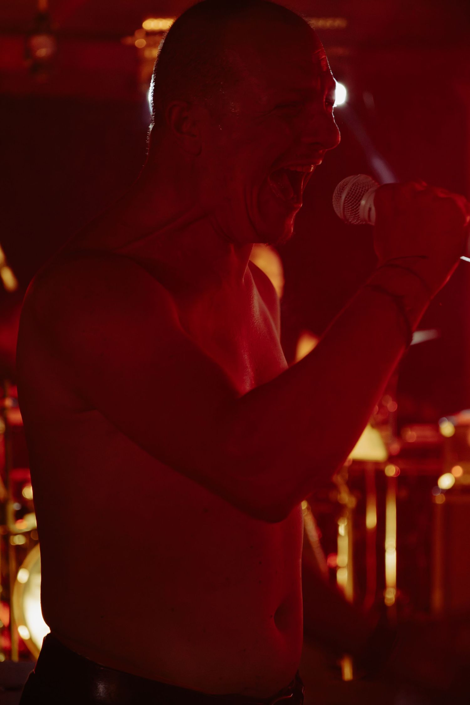 Shirtless male vocalist performs intensely on stage under deep red concert lighting.