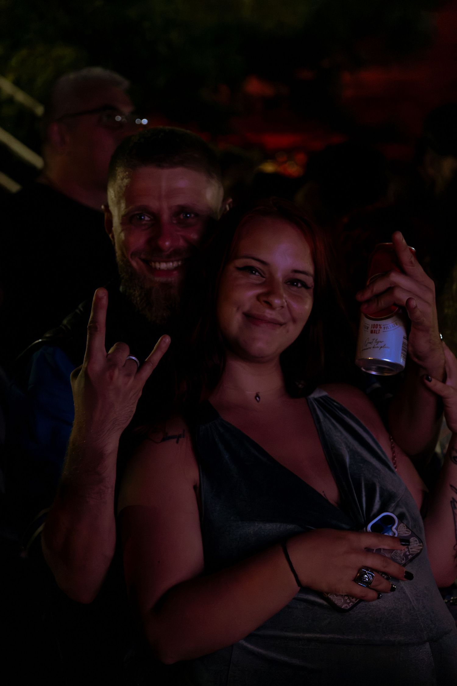 Smiling couple at rock concert, man making devil horns gesture, woman holding beer bottle.